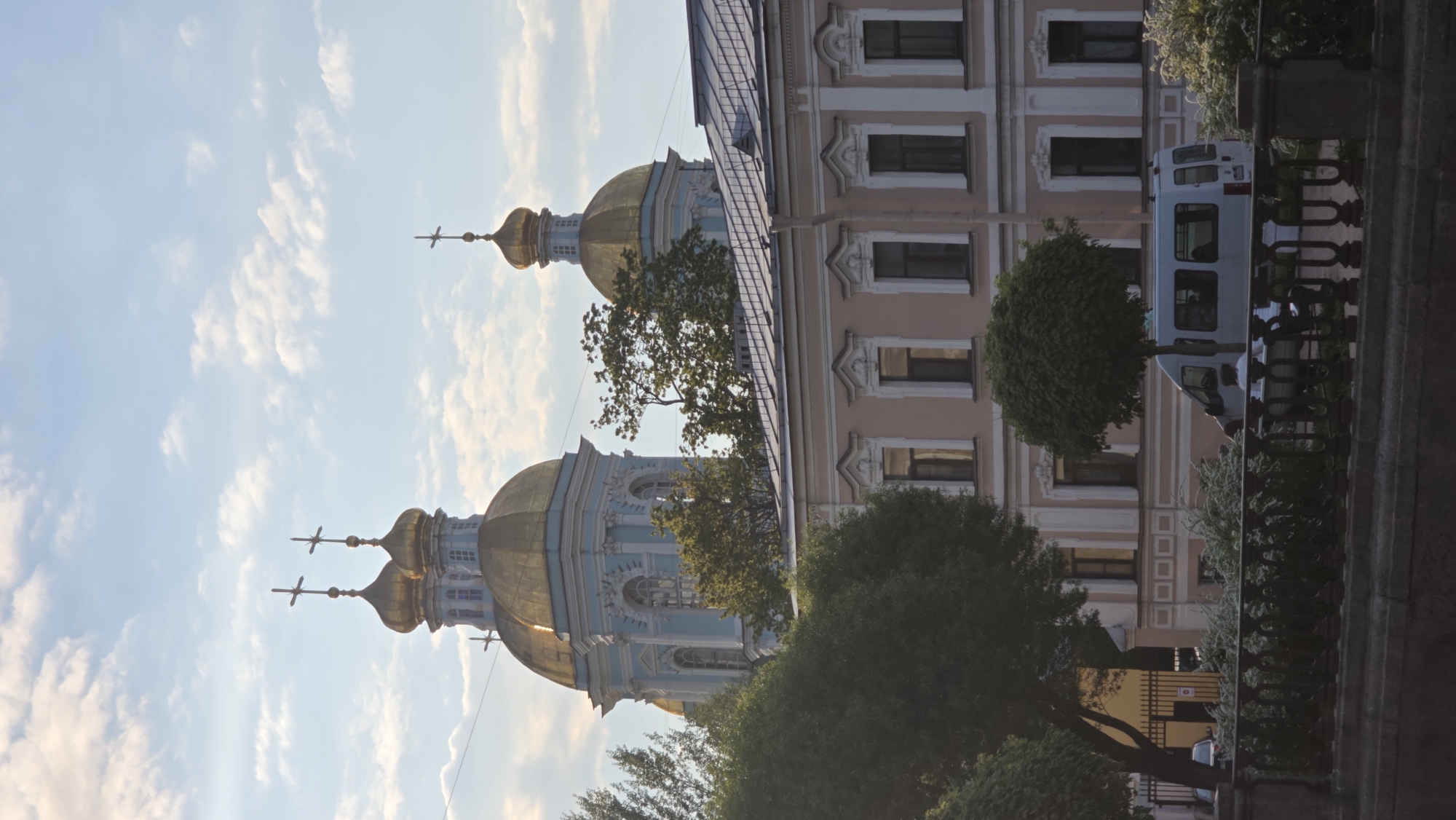 The image shows a view of the Saint Nicholas Naval Cathedral in Saint Petersburg, Russia. The cathedral features multiple golden domes topped with crosses. Part of a building with a light pink facade and windows is visible to the right. A van is parked adjacent to the building. Trees are present, partially obscuring the structures. The sky is light blue with scattered white clouds.