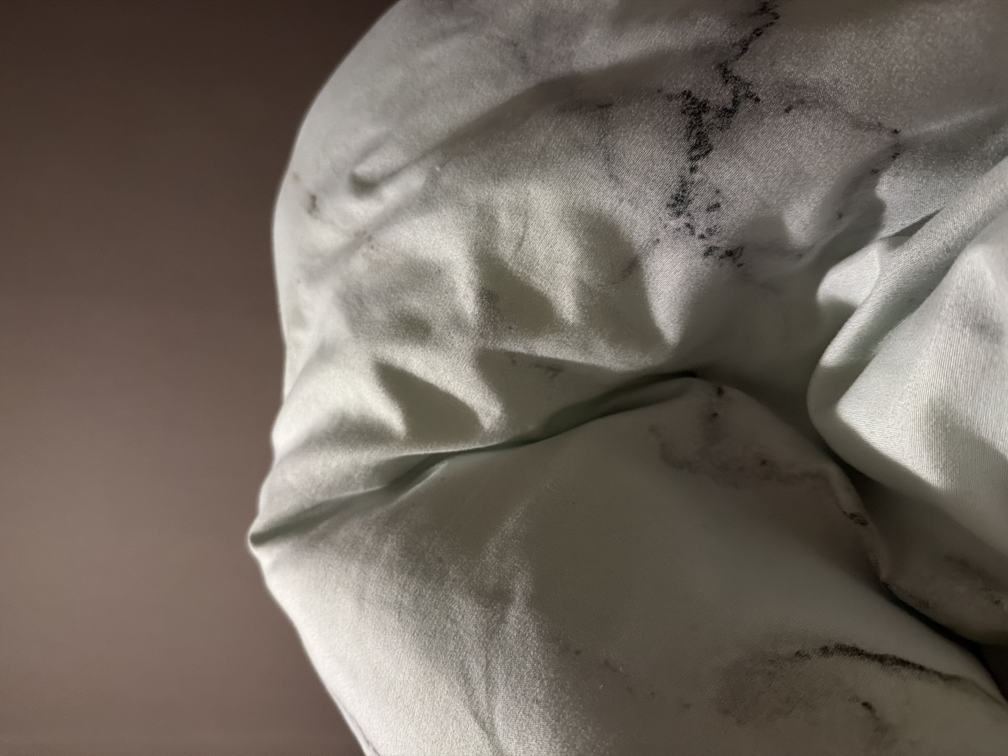 The image is a close-up, low-angle shot of crumpled, light green fabric, possibly a duvet or comforter, against a blurred brown background. The fabric is wrinkled, creating shadows and highlights that emphasize its texture. Dark, irregular smudges and stains are visible on the fabric, particularly in the lower right and center portions of the image. These stains appear to be a dark gray or black color and vary in size and density. The lighting is dim, with a light source likely coming from above and slightly to the left, casting shadows that deepen the folds of the fabric. The overall impression is one of disarray and possibly uncleanliness due to the visible stains. No specific location in a city is identifiable.