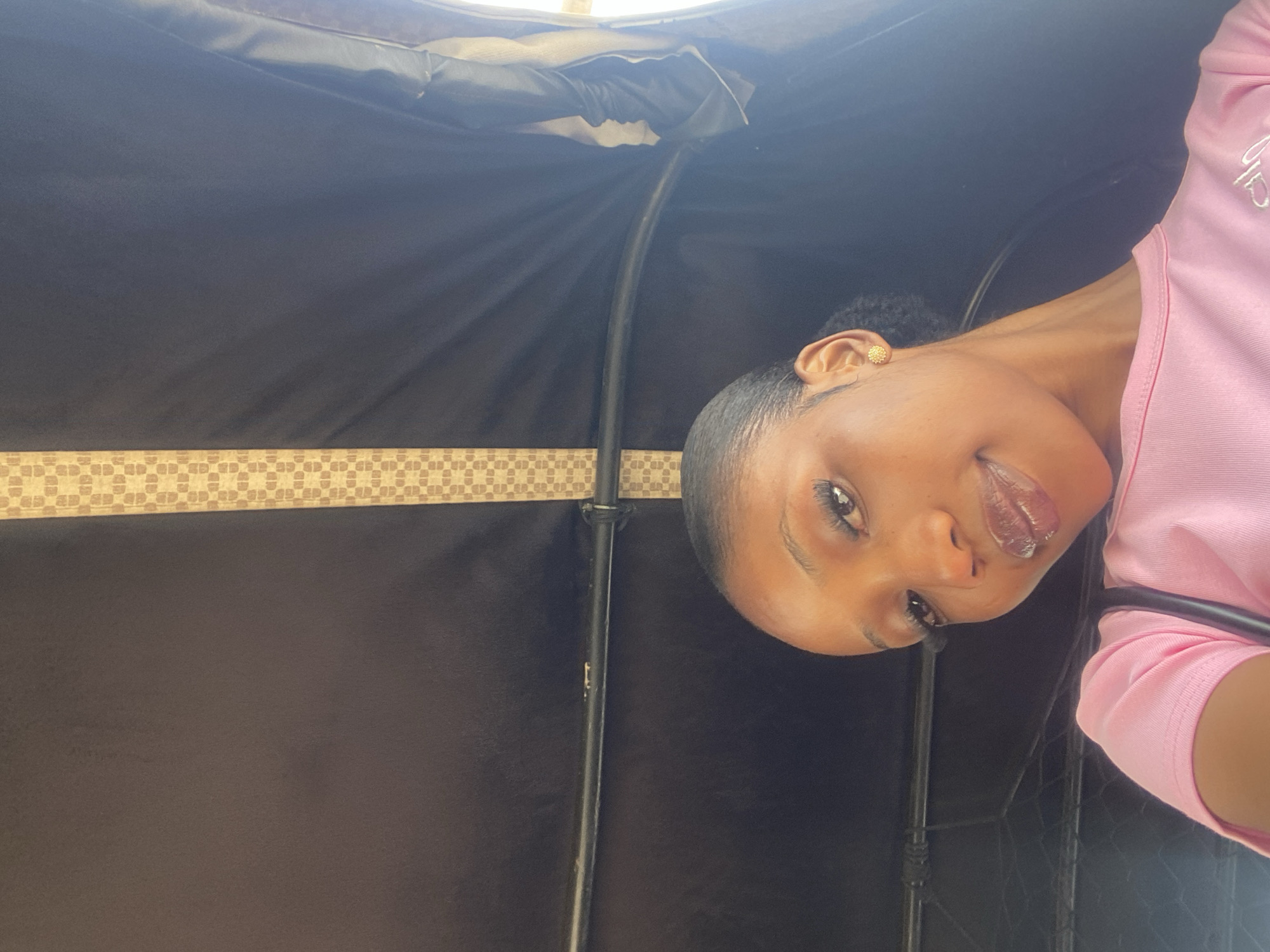 A young Black woman with dark hair pulled back into a ponytail and wearing a pink t-shirt is looking into the camera. She has on subtle makeup, including mascara and lip gloss, and a gold earring in her left ear. She is inside what appears to be a vehicle, possibly a trishaw, given the black fabric covering the interior and the curved metal bar visible in the foreground. The background is dark, with a strip of light brown patterned fabric running vertically down the center and a sliver of bright light on the right side, suggesting an open window or exit. The image was taken in Barakin Lerawi, Nigeria.