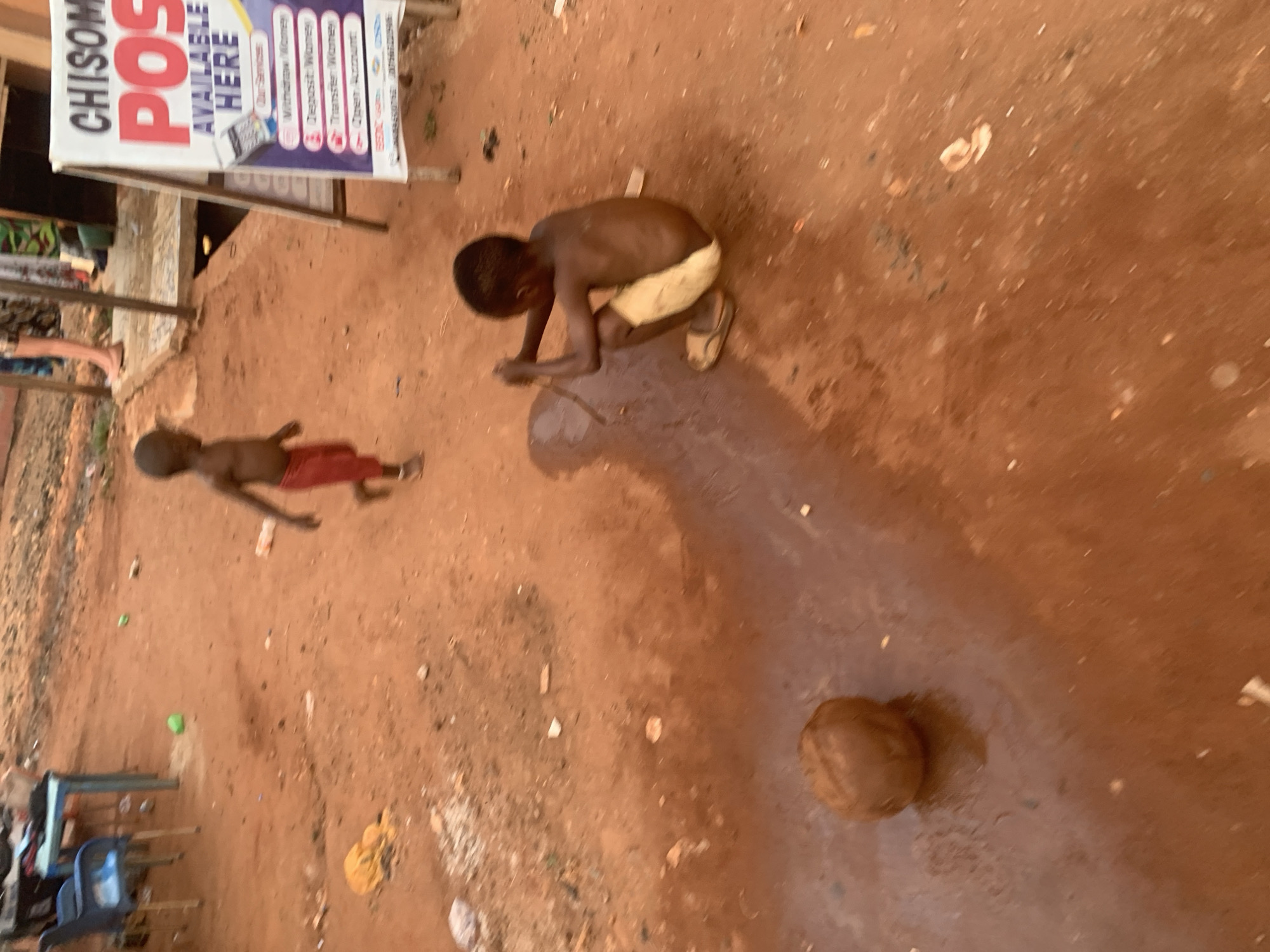 Two young children are playing on a dirt ground in Ukwu-Abwa, Nigeria. One child is shirtless, wearing light-colored shorts and sandals, crouching and holding a stick. The other child is shirtless, wearing red shorts, standing. A coconut lies on the ground nearby. A sign advertising banking services is visible in the background. Chairs and other items are near the sign.