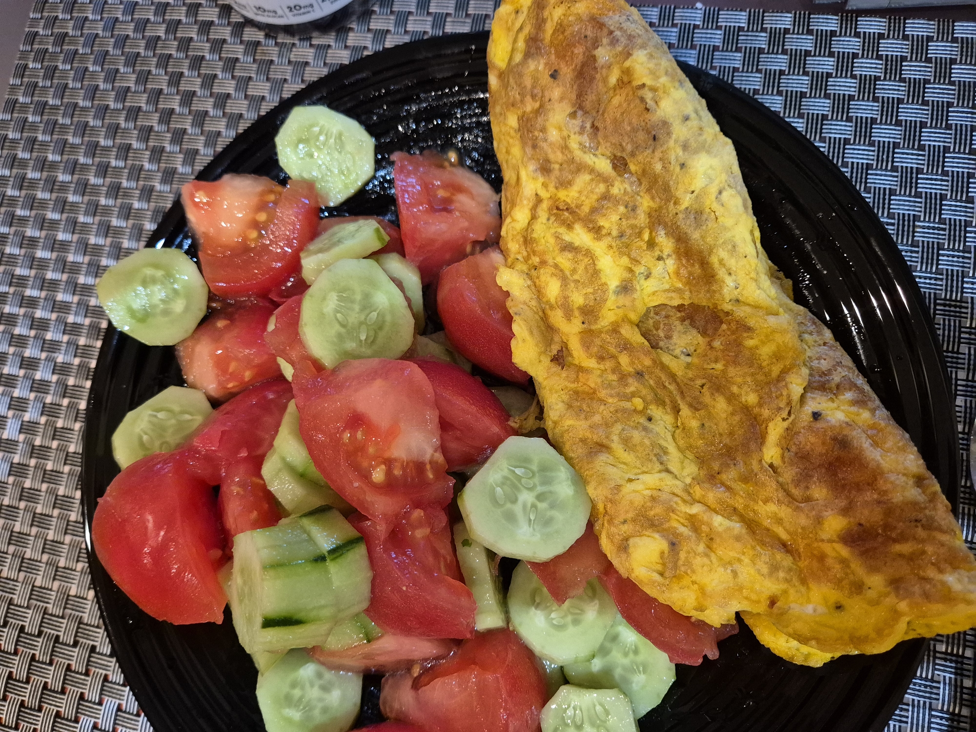 A close-up, overhead shot shows a black plate filled with a meal. The meal consists of a chopped tomato and cucumber salad and a folded omelet. The salad is composed of bright red tomato wedges and pale green cucumber slices, some of which show the seeds. The omelet, golden yellow and slightly browned in places, is folded over on itself. The plate sits on a textured placemat with a woven pattern of dark grey and white. The background is blurred, but appears to be a neutral surface.