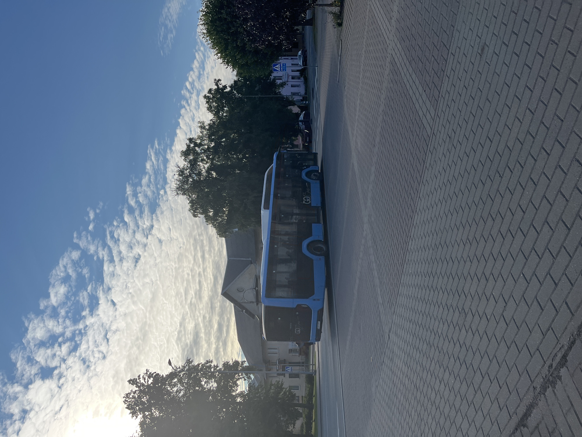A blue bus is driving down a street in Kohtla-Järve linn, Estonia. The sky is blue with white clouds. Trees are on the side of the street, and there are buildings in the background. The street is paved with gray bricks.