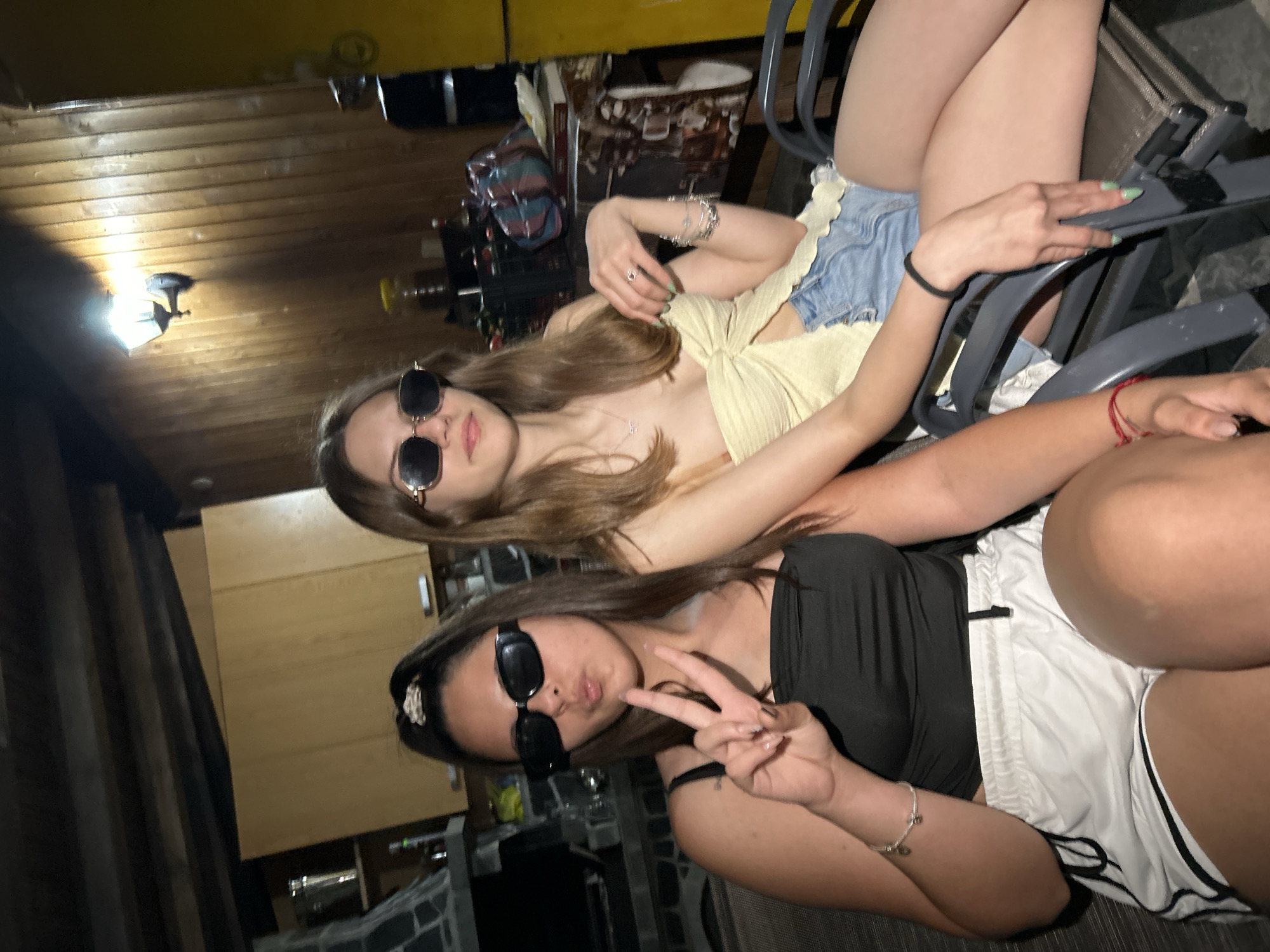Two young adult females are depicted seated indoors in an unknown location. The lighting is dim, with a prominent warm light source visible on the right wall.
The individual on the left has dark brown hair styled in a high ponytail secured with a patterned scrunchie. She wears rectangular black sunglasses, a black strapless top, and white athletic shorts with black trim. Her left hand is raised, displaying a "peace" sign with her index and middle fingers, while her lips are puckered. A silver charm bracelet is visible on her right wrist.
The individual on the right has long, light brown hair parted in the middle. She wears hexagonal gold-rimmed sunglasses, a light yellow or cream-colored strapless top tied in the front, and light blue denim shorts. Her left hand rests on her lap, adorned with multiple rings, including one with a red stone, and several silver bracelets. Her fingernails are painted light green. Her right hand rests on the armrest of her chair, with a black elastic hair tie on her wrist. She exhibits a subtle smile.
Both subjects are seated in dark-colored, possibly metal or plastic, chairs.
The background on the left features a stone or tile-clad structure, potentially a fireplace or oven, with a metal bucket on its surface. Above this, light brown wooden cabinets are visible. The ceiling exhibits exposed dark wooden beams.
The background on the right consists of vertical wooden wall paneling. A wall-mounted light fixture with a glowing bulb illuminates this section of the wall. Various objects are discernible on shelves and surfaces, including a yellow container, items within crates, and a stack of books. A portion of a yellow appliance, possibly a refrigerator or cabinet, is visible on the far right.