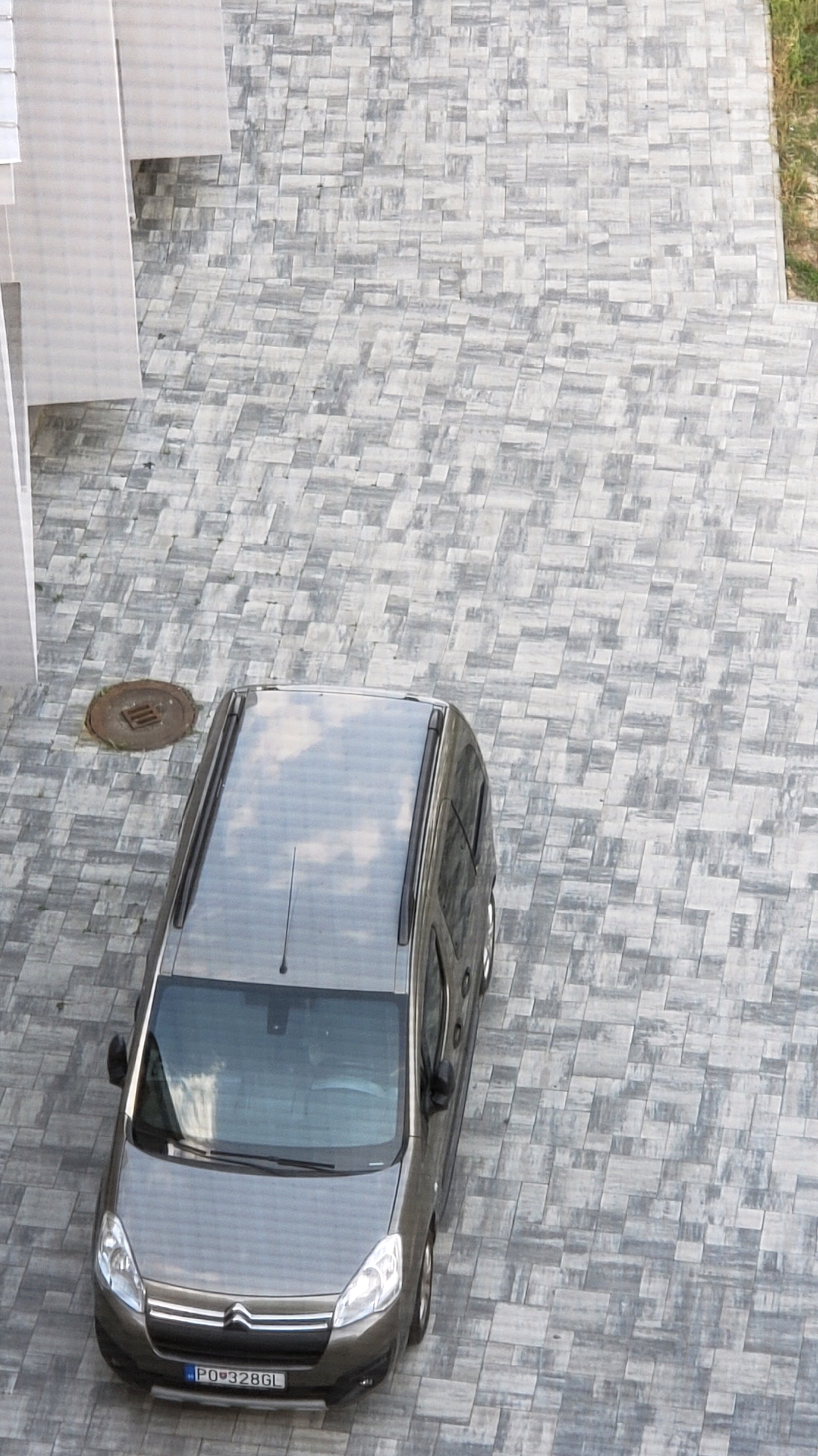 An aerial shot captures a gray Citroen Berlingo parked on a paved driveway made of gray interlocking stones in Prešov, Slovakia. The car's license plate reads "PO 328GL." The driveway is bordered by a patch of green grass on the right side and a modern building with light-colored walls and vertical accents on the left. A circular metal grate cover is visible on the pavement near the car. The sky, reflected in the car's roof, shows a scattering of white clouds.