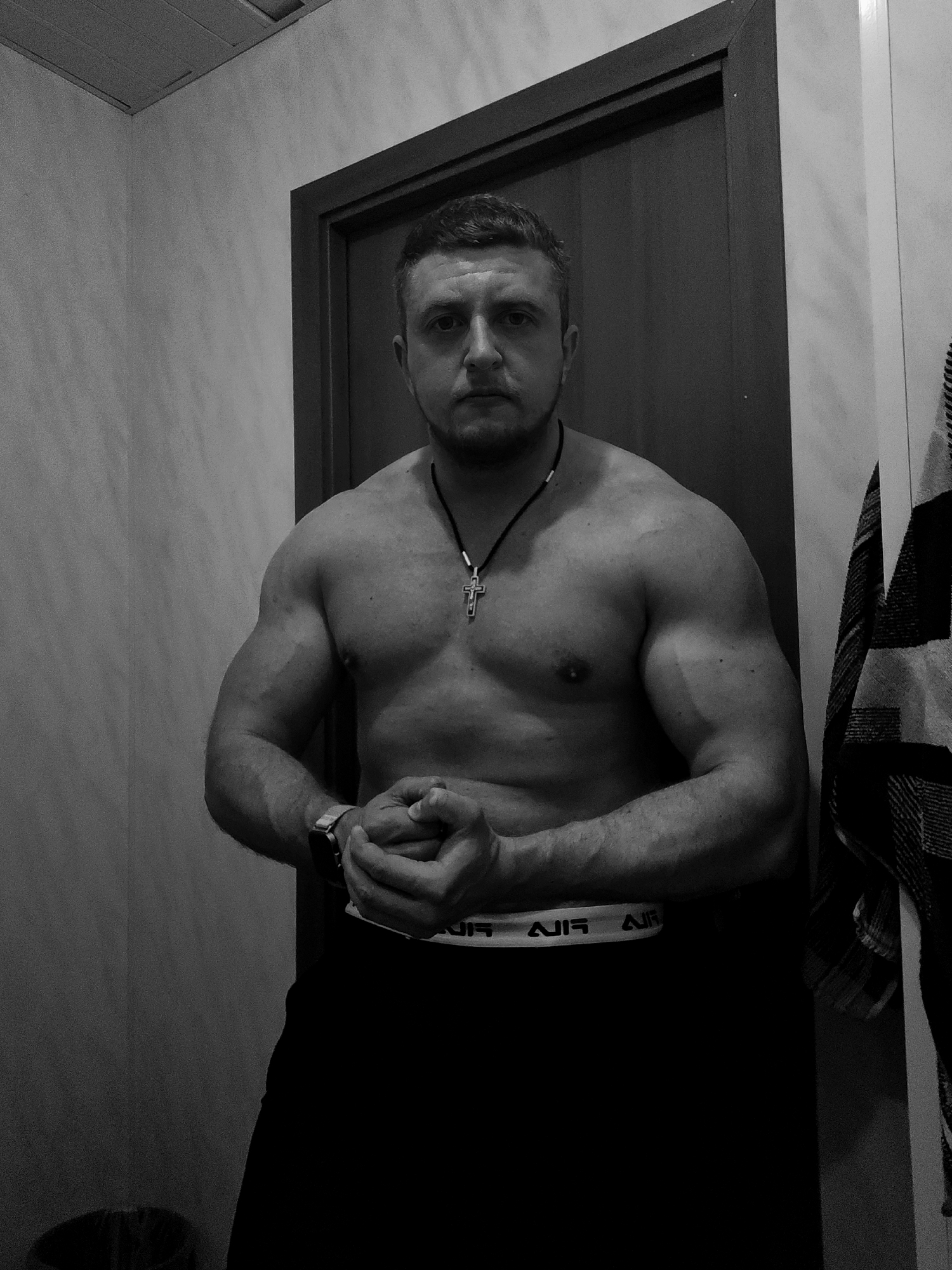 A black and white medium shot photograph depicts a muscular man posing in what appears to be a bathroom. The location is Unknown, Unknown. The man has short, dark hair and a beard. He is shirtless, wearing a necklace with a cross pendant, black pants with the word "AIR" repeated on the waistband, and a watch on his left wrist. His hands are clasped together in front of him. Behind him is a closed door with a dark frame. To the left is a wall and to the right a checkered towel.