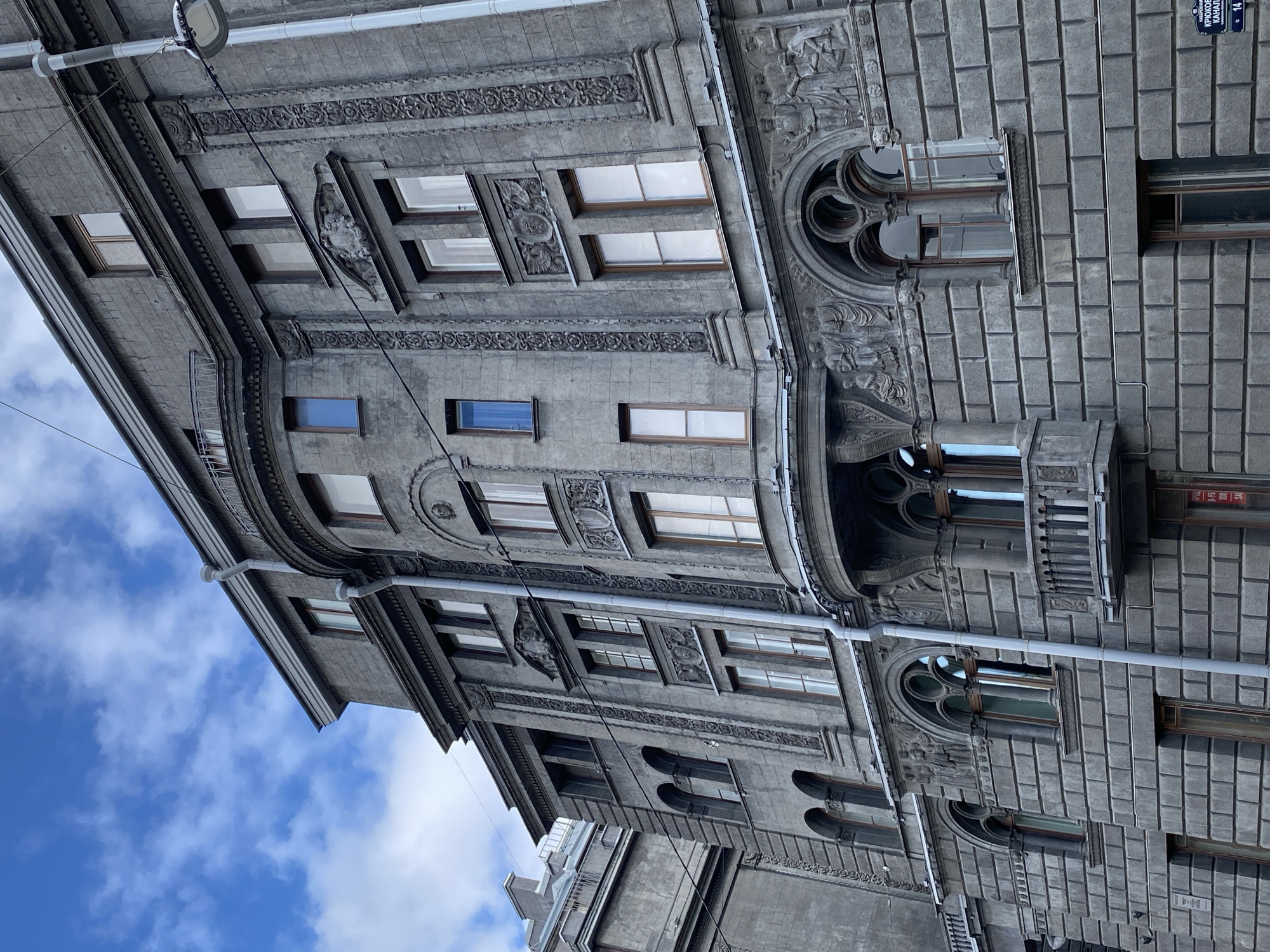 This is a low-angle, exterior, eye-level shot of a large, ornate, grey stone building in Saint Petersburg, Russia, under a blue sky with scattered clouds. The building features intricate architectural details, including decorative carvings, columns, arched windows with decorative tracery, and balconies. A downspout runs vertically along the facade. To the right, a street lamp on a pole is visible. In the lower right corner, a street sign with Cyrillic text and numbers "14" and "13" is affixed to the building. The building appears to be multi-story, with many windows of various sizes.