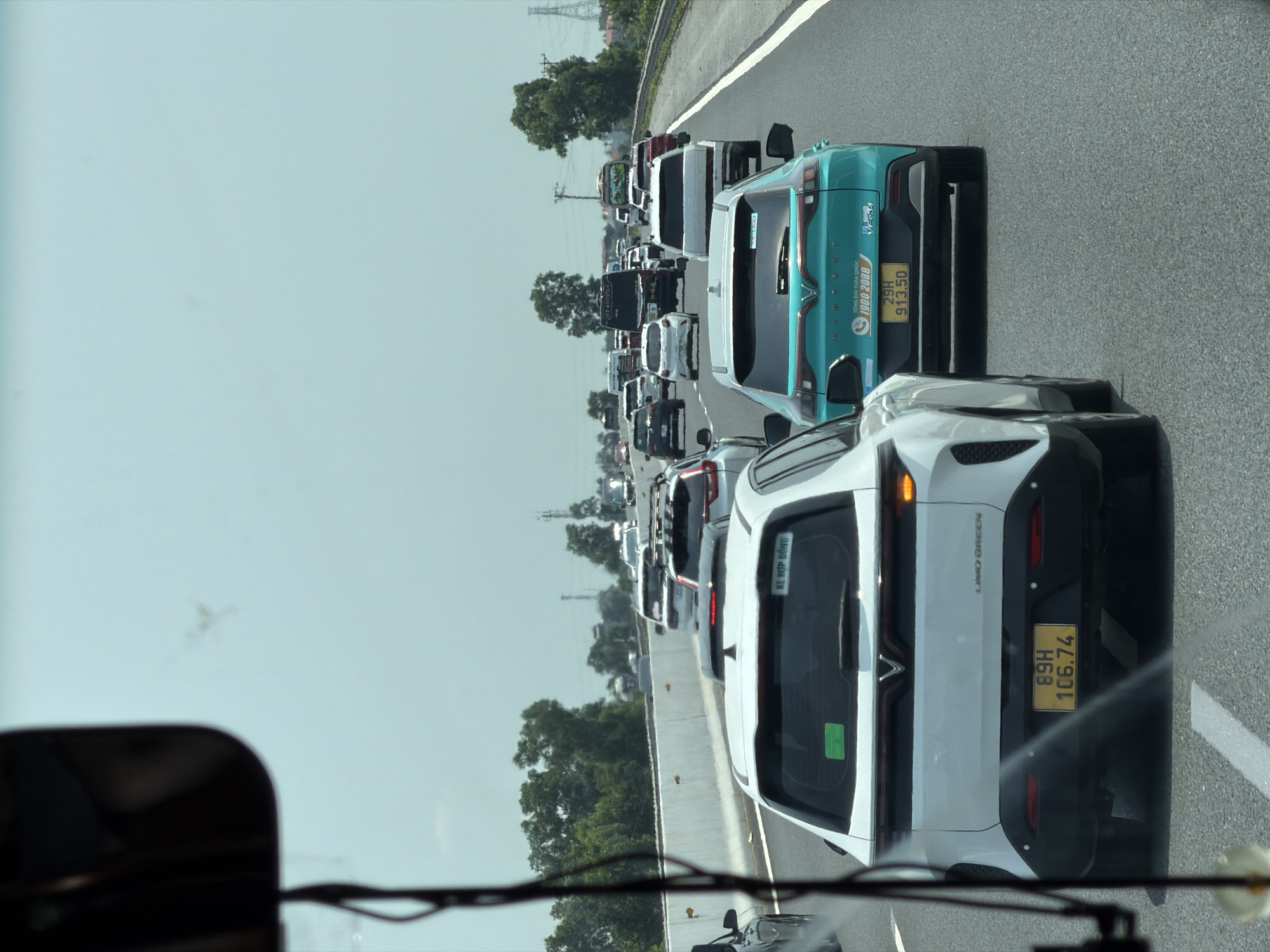 The image shows a traffic jam on a highway, likely in Hải Phòng, Vietnam. The scene is observed from within a vehicle, looking forward through the windshield. A white LIMO GREEN car with license plate 89H-106.74 is directly in front, followed by a turquoise VINFAST VF-e34, a brand known in Vietnam. This VINFAST has the license plate 29H-913.50 and advertises a taxi service with the phone number 1900 2088. The congested road stretches into the distance with numerous cars and trucks. Trees line the side of the road, and power lines are visible in the background. The sky is overcast.