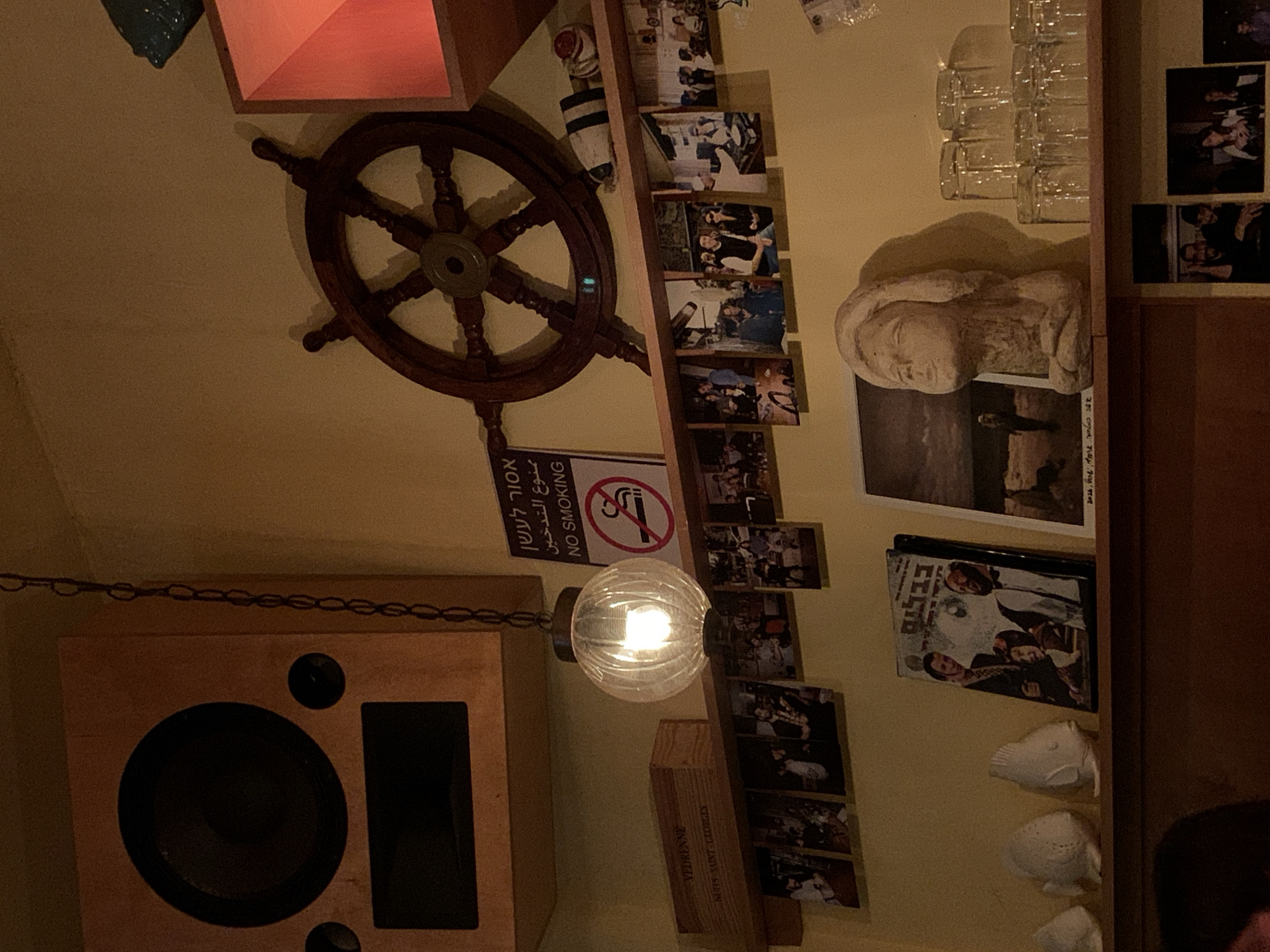 This image depicts the interior of an establishment in Tel-Aviv, Israel. The wall is painted a uniform beige color and is adorned with various objects under dim, warm artificial lighting.
In the upper left quadrant, a large, rectangular wooden speaker cabinet, featuring a central circular speaker and smaller components, is mounted. A spherical, ribbed glass pendant light fixture, illuminated and emitting a warm glow, hangs from a metal chain in the upper-center of the frame.
To the upper right, a dark wooden ship's wheel is affixed to the wall. Adjacent to it, a rectangular "NO SMOKING" sign is displayed, featuring text in Hebrew, Arabic, and English, along with a universal no-smoking symbol (a cigarette with a diagonal line through it). A decorative object resembling a striped marine fender or buoy rests on a shelf above. A reddish, geometric light fixture or shelf element is also visible in the upper right corner.
A horizontal wooden shelf spans the mid-section of the wall. On this shelf, a wooden box with the label "VEDRENNE NUITS SAINT-GEORGES" is placed. Directly below this shelf, a row of approximately twelve unframed photographs are affixed to the wall, depicting various individuals in social gatherings and candid group settings.
Below the first shelf, a second wooden shelf extends across the lower portion of the frame. From left to right on this shelf: three white, stylized ceramic fish sculptures are displayed. Next to them, a framed artwork or poster features two men and a child, with the Hebrew word "בבלעם" visible above their heads. A photograph depicting a single figure on a beach is positioned beside this artwork. Further to the right, a light-colored, possibly plaster or stone, bust of a human head with closed eyes stands upright. To the far right, multiple clear glass tumblers are stacked in two rows. Below this lower shelf, several more unframed photographs are visible, showing additional people in social environments. No active individuals are present within the primary frame of the image.