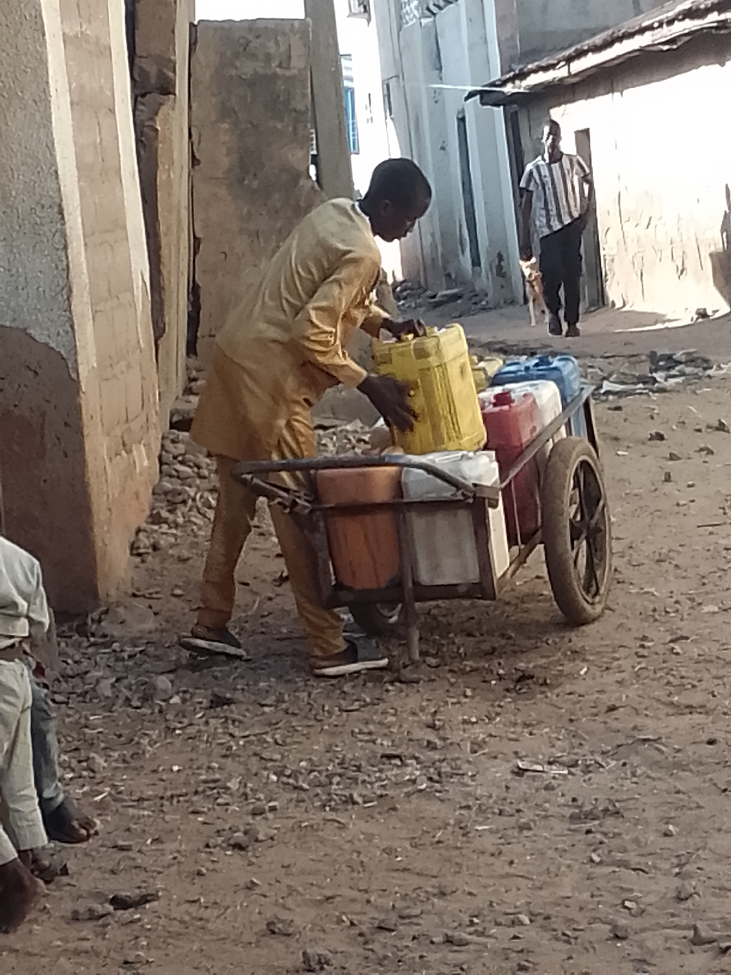 In the city of Unknown, Unknown, a person in a tan outfit pushes a cart laden with yellow, red, and blue plastic containers. The cart has a metal frame and two wheels. The person's left hand rests on the yellow container. In the background, another person wearing a striped shirt and black pants walks away from the viewer, accompanied by a small dog. The scene takes place on a dirt path, with buildings made of concrete and corrugated metal lining the sides. The buildings appear to be in a state of disrepair. The lighting suggests it is daytime, with shadows cast on the ground.