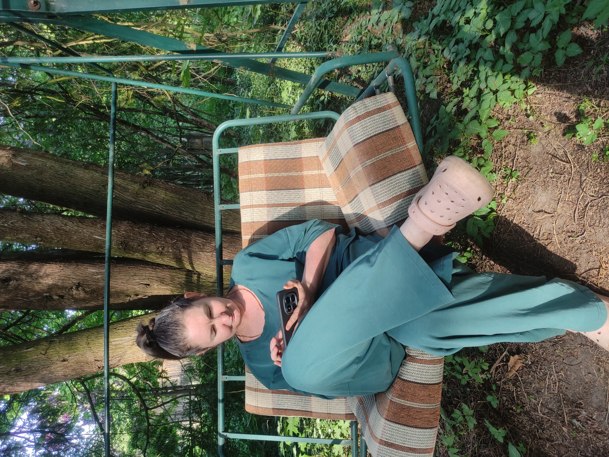 A woman is sitting on a porch swing in a wooded area. She is wearing a teal outfit and pink Crocs. She has dark hair pulled back in a bun. She is holding a black cell phone in her hands. The porch swing has brown and beige striped cushions. The background is filled with trees and greenery. The location is impossible to ascertain based solely on the visual information provided.