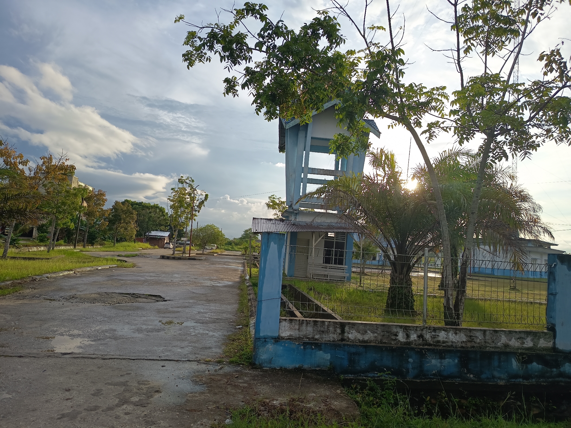 The image depicts an outdoor scene in Singkil, Indonesia, featuring a weathered concrete road or parking lot in the foreground with visible potholes and puddles. To the right, a blue and white structure, possibly a guardhouse or small office building, is partially obscured by a metal fence and overgrown vegetation. A tall, blue and white tower-like structure stands next to the building, partially covered by tree branches. The background shows more trees, a low-lying building with a corrugated metal roof, and distant vehicles. The sky is partly cloudy with sunlight breaking through, suggesting it is either early morning or late afternoon. The environment appears somewhat unkempt, with tall grass growing around the structures and along the roadside.