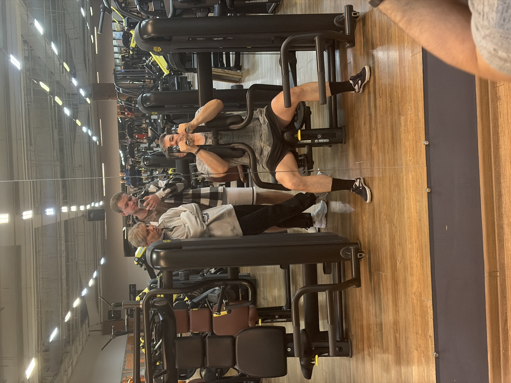 A photograph shows a gymnasium interior with various exercise machines. In the foreground, a man is seated on an exercise machine, his legs positioned under the machine's arm with both his hands holding on to its handles. A person in a plaid shirt is standing, taking a picture of him. Nearby, another person can be seen lying down, with their feet toward the edge of the frame and wearing white sneakers. The reflection from a mirror occupies the right side of the image, partially obscuring the view. The gymnasium appears to be in Chaikovsky, Russia.
