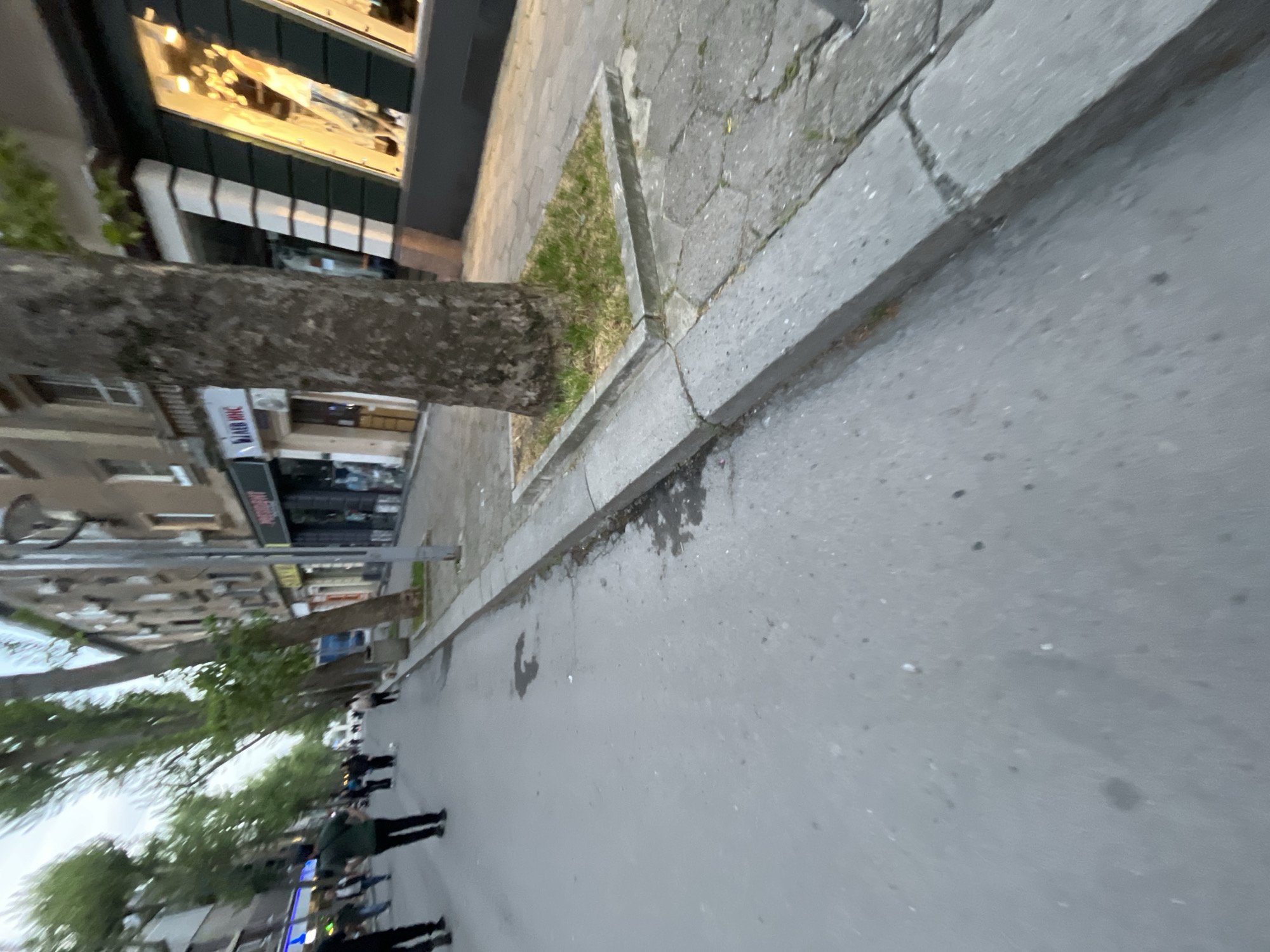 The image shows a street scene in Burgas, Bulgaria. People are walking on the sidewalk. There are buildings and trees along the street. The photograph is taken at street level.