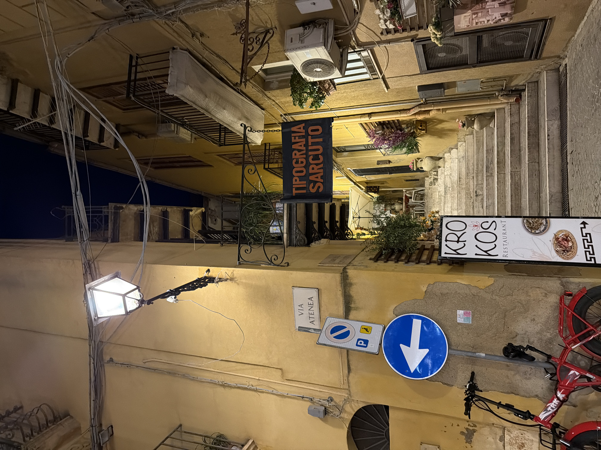The image depicts a narrow street in Agrigento, Italy, at night. The buildings are old and yellow, with balconies and air conditioning units visible. A street lamp illuminates the scene, casting long shadows. There are several signs on the wall, including one for "VIA ATENEA" and another for "TIPOGRAFIA SARCUTO." A red bicycle is parked on the left side of the street, and a sign for "KRO KOS RESTAURANT" is visible on the right. The street slopes upwards, with a set of stairs leading into the distance. The overall atmosphere is dimly lit and somewhat mysterious, suggesting a quiet evening in a historic Italian city.