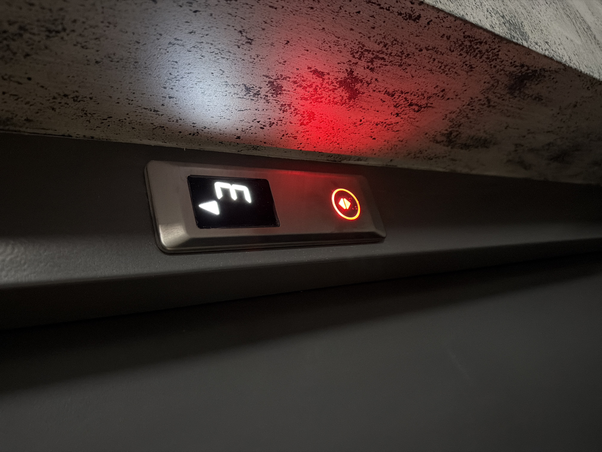 The media file shows a close-up of an elevator control panel. The panel is mounted on a dark grey surface, and above it, a textured ceiling is visible, with areas illuminated by white and red light.
The primary object of focus is the elevator button. It is a rectangular button with a silver-colored metal frame. Inside the frame, there are two illuminated elements. On the left is a black rectangle displaying a white symbol that resembles the letter "M" with an upward-pointing arrow below it. On the right is a circular button illuminated with a red-orange light. Inside the circle, a symbol of an upward-pointing arrow is visible.
The lighting suggests an indoor setting, possibly a building in Plovdiv, Bulgaria. The red light reflecting on the ceiling might indicate the elevator is on a specific floor or in a particular operational state. There is no visible text other than the illuminated symbols on the button. The image captures a static moment, with no activity or people depicted. The overall impression is one of functional technology within an architectural space.
