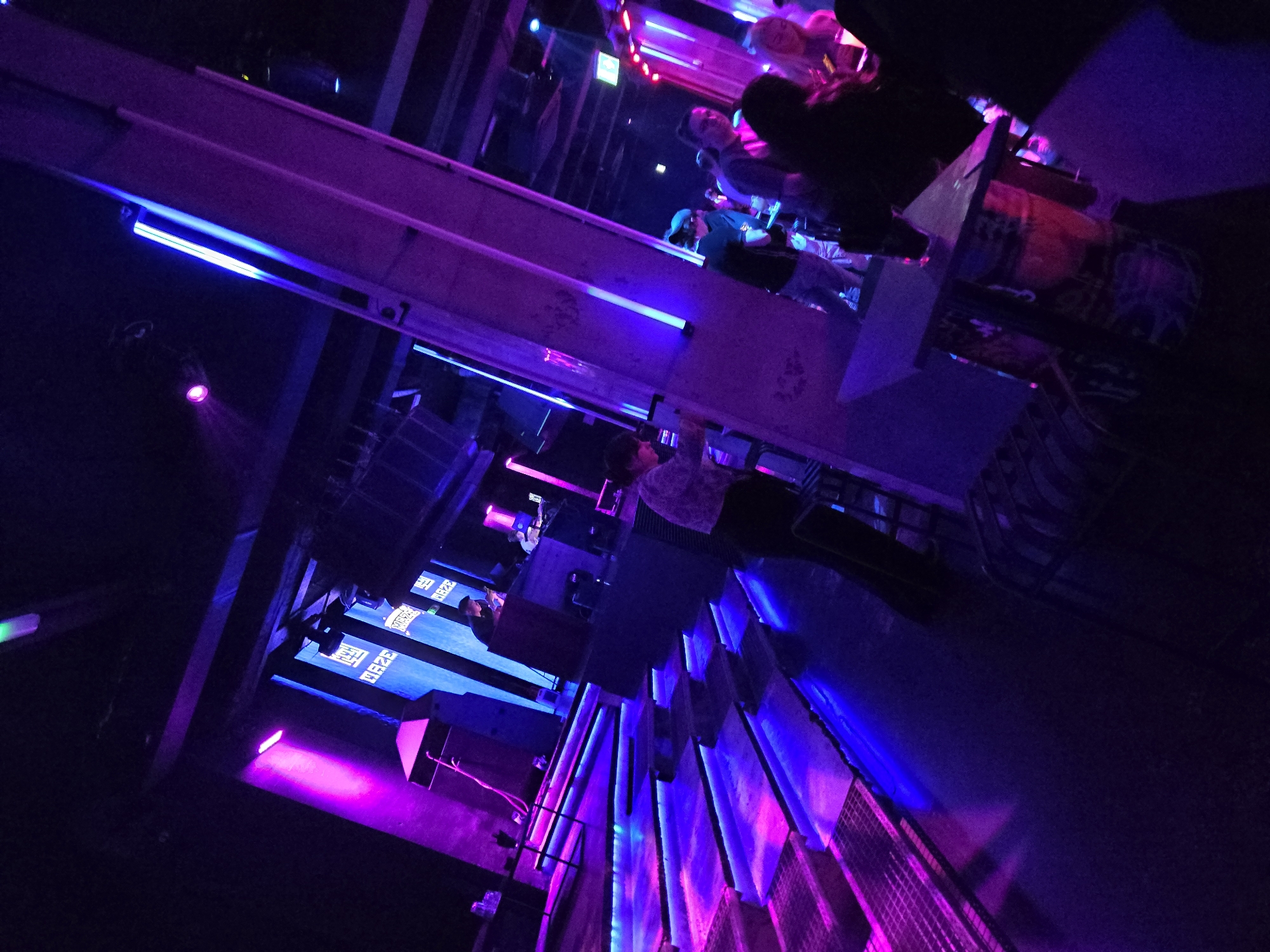 The image depicts the interior of PLAN B, a bar in Varna, Bulgaria. The scene is dominated by blue and purple lighting, creating a nightclub atmosphere. There are several people present, some standing and others seated. A bar area is visible with bottles and glasses. The architecture features linear lighting fixtures and geometric designs. There are some shadows present.