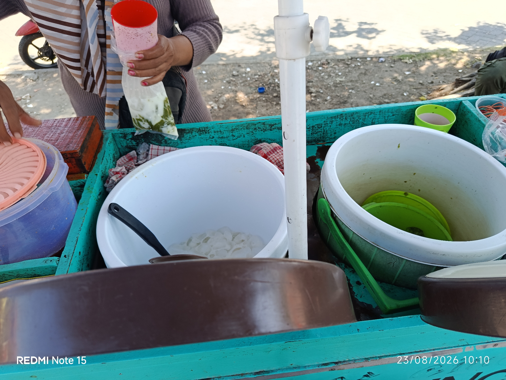 The image shows a close-up view of a street vendor's cart in Keboansikep, Indonesia, on August 23, 2026, at 10:10 AM. The cart is painted turquoise and appears to be made of wood.
In the foreground, a large, dark brown bowl or container is visible. To the left, a pink plastic lid sits on top of a translucent purple container.
The middle section of the cart holds several items. A large white bowl contains what appears to be ice cubes. A black ladle rests inside this bowl. Next to it, another large white container holds a green plastic lid. A green ladle-like object is positioned within this container, partially submerged. A small green cup containing a light pink liquid is also visible. To the right of the green cup, a clear plastic bag filled with a white and green mixture is held by a person's hands, who is also holding a red-topped cup. The person is wearing a striped scarf and a brown sweater. A red and white checkered cloth is placed on the side of the cart.
In the background, a red motorcycle is parked, with its rear wheel visible. The ground is covered with small stones and debris. The lighting suggests it is daytime, with shadows cast by what appears to be foliage overhead. The overall scene depicts a typical street-side food or beverage stall.