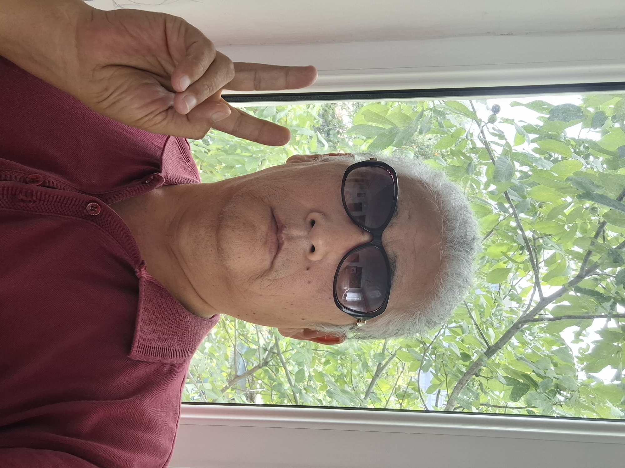 A middle-aged East Asian man with grey hair is posing for a selfie in front of a window with green foliage visible outside. He is wearing dark sunglasses and a maroon polo shirt. His left hand is raised, with his index and middle fingers extended in a peace sign. The lighting suggests it is daytime, possibly indoors with natural light from the window. The background is blurry, with green leaves and branches of a tree. The man's skin tone is tan, and his face appears weathered. The overall impression is a casual, personal photograph. The image does not contain any information that would allow for its localization in Almaty, Kazakhstan.