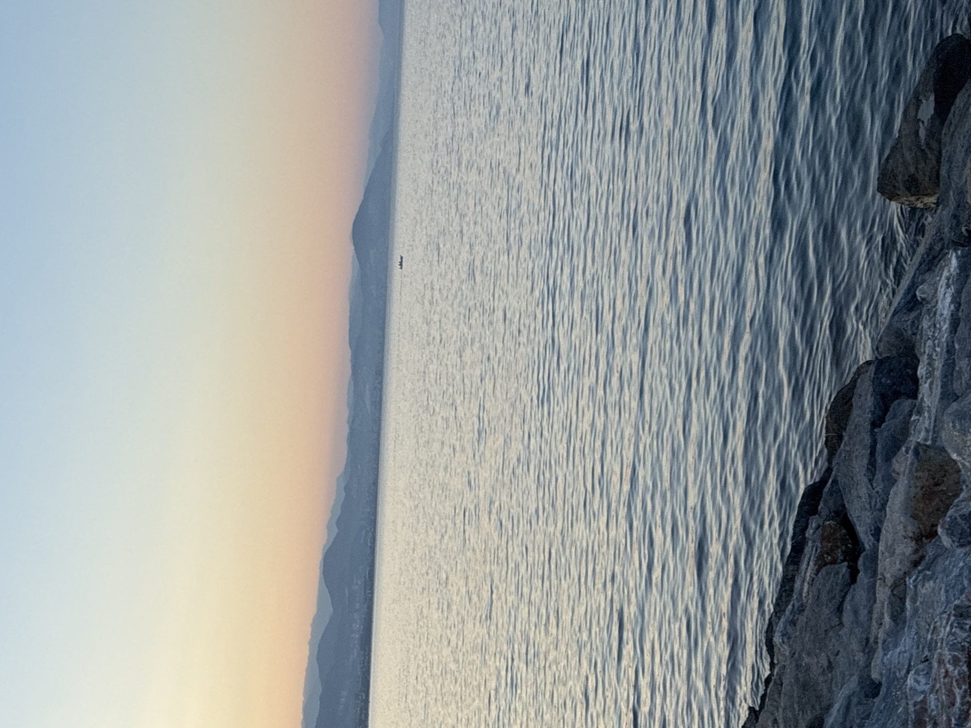 An outdoor, eye-level, wide shot captures the calm sea at sunset. The water's surface is rippled, with sunlight reflecting off it. In the distance, a line of mountains forms the horizon, with a faint city visible along the coastline. The sky displays a gradient of soft hues, transitioning from a pale orange near the horizon to a clear blue overhead. In the middle of the water, a small boat with what appear to be three people is visible. The foreground is occupied by large, grey, weathered rocks, suggesting a waterfront promenade or breakwater. The overall scene evokes a peaceful and tranquil atmosphere. Based on the mountainous coastal landscape and the appearance of the water and sky, this scene is likely located in Alanya, Turkey.