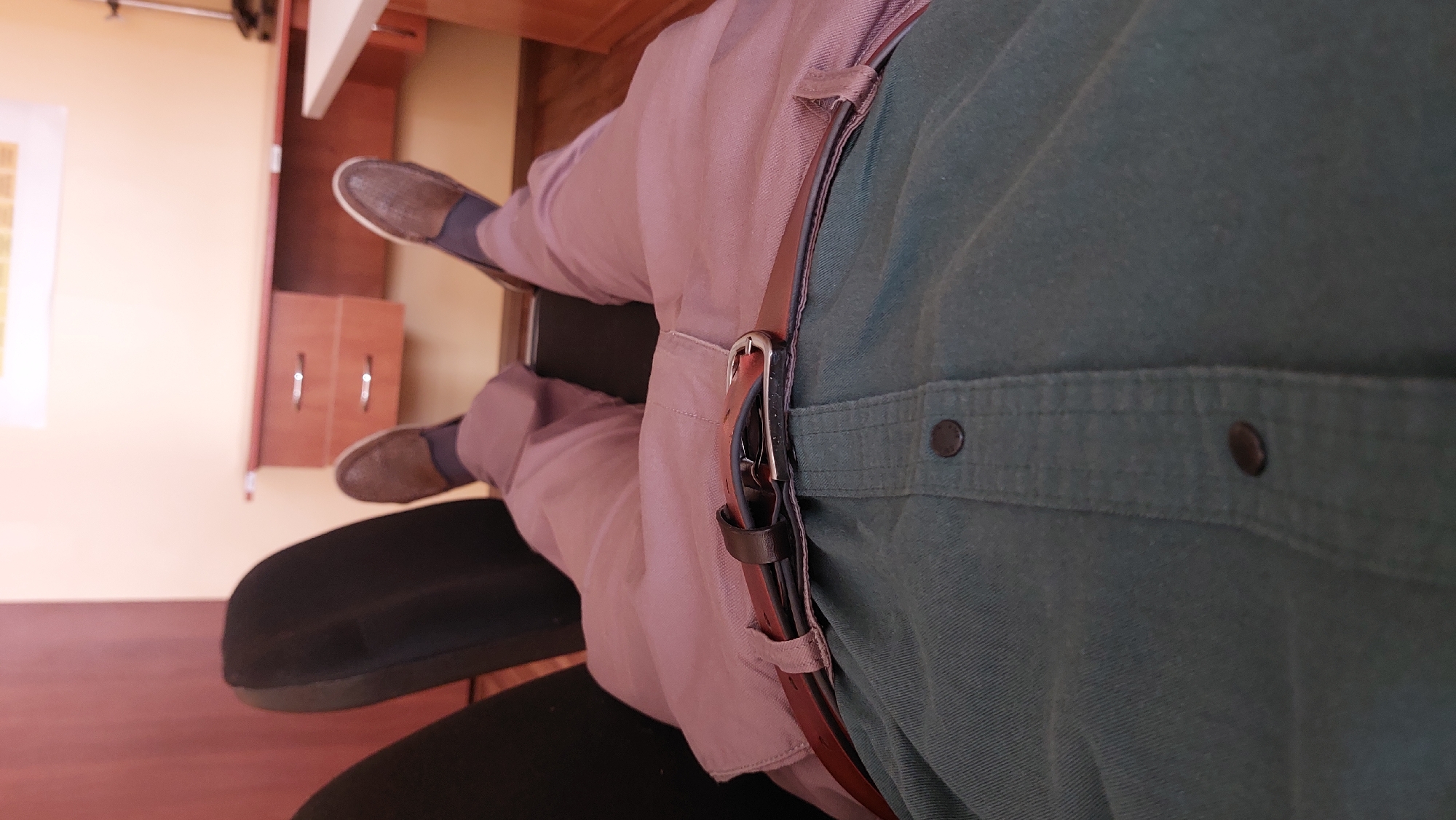 A person wearing a green button-down shirt and mauve pants sits on a black office chair. They are wearing brown slip-on shoes. A brown leather belt with a silver buckle is visible around their waist. The scene appears to be inside an office, with wooden furniture and a light yellow wall visible in the background. The photograph is oriented vertically.