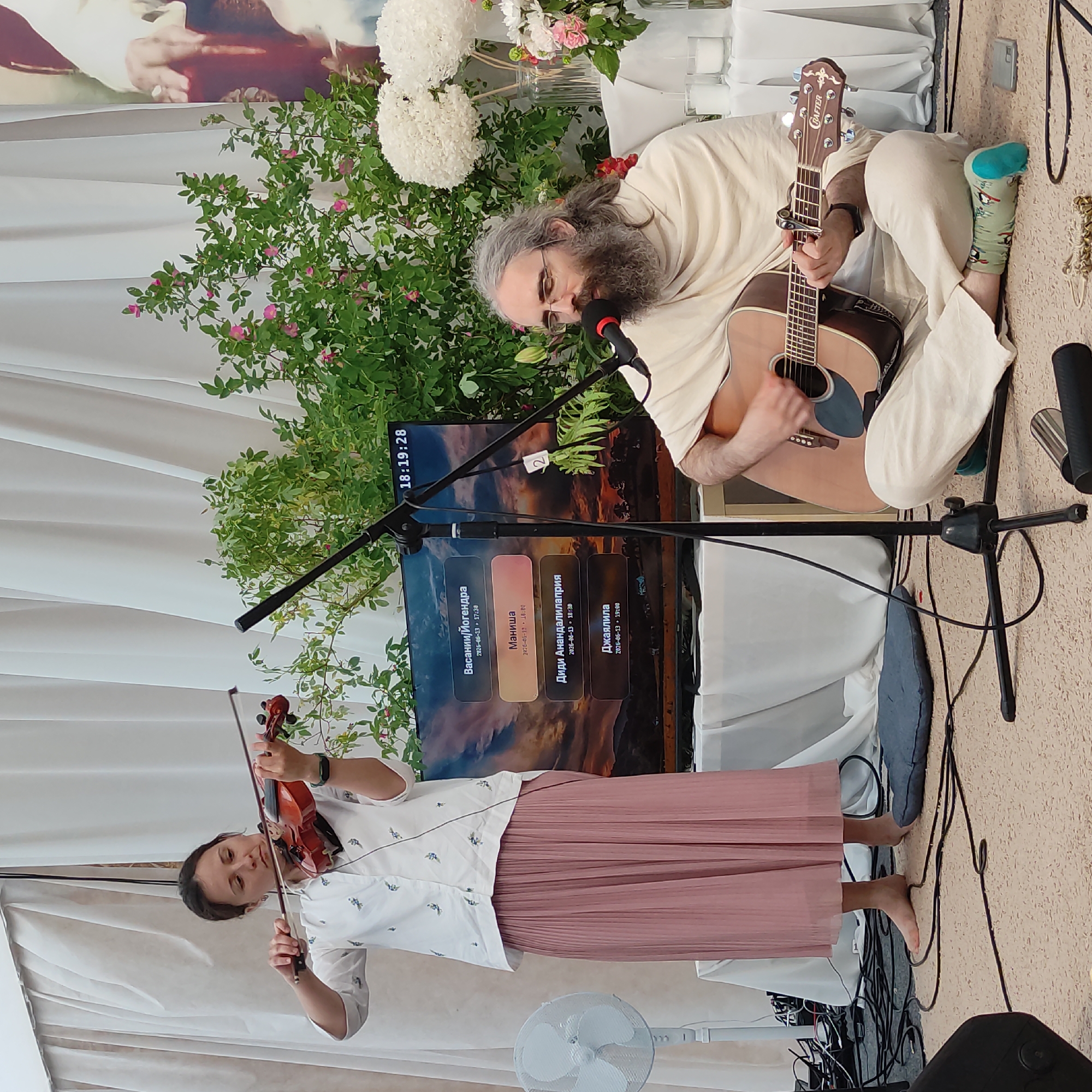 In the city of Zhirnosovo, Russia, at an outdoor event held in what appears to be a tent or covered space with draped white fabric walls, a woman stands to the left playing a violin, while a man sits to the right playing an acoustic guitar and singing into a microphone. The woman is wearing a white, short-sleeved shirt with a floral pattern and a long, pleated pink skirt. She holds a brown violin under her chin, bowing with her right hand and fingering the strings with her left. The man has long grey hair and a beard, and wears glasses. He is dressed in a cream-colored robe and is seated on the floor, his legs crossed. He holds a light brown acoustic guitar, strumming with his right hand and fretting with his left. A black microphone is positioned in front of him. Behind the performers is a large screen displaying a sunset landscape with text in a Cyrillic language, indicating times and names. Various plants, including a rose bush with pink flowers, are visible around the screen and to the right of the man. White flowers in vases and lit candles are also present on a table to the right. Electrical cords and equipment, including a fan on the far left, are scattered on the carpeted floor, suggesting a stage setup. The overall scene depicts a musical performance in a serene, perhaps spiritual or cultural, setting.