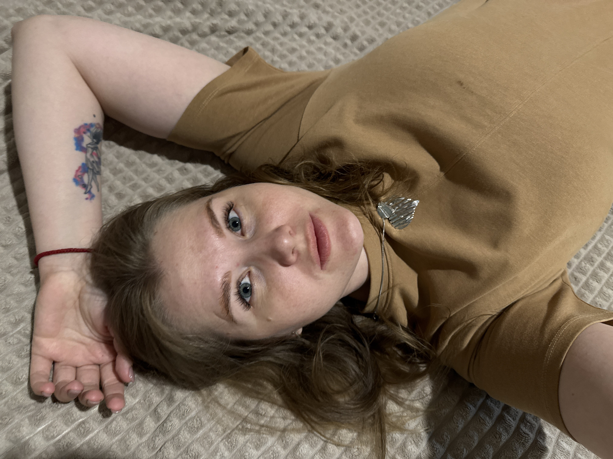 A young woman is captured lying on her back on a textured, light brown or beige blanket or bedspread, featuring a waffle-like pattern. Her head is tilted to her right, and she looks directly at the camera with blue eyes and a neutral to calm expression. She has fair skin, visible freckles, and long, wavy brown hair that is spread around her head.
She is wearing a plain, short-sleeved, light brown or camel-colored t-shirt. A silver-colored pendant, possibly a stylized heart or geometric shape, hangs on a thin chain around her neck. On her right wrist, she wears a thin red string bracelet. A prominent tattoo is visible on her upper left arm, depicting an abstract figure, possibly an astronaut or a person in motion, surrounded by watercolor-style splashes of blue, pink, and red. Her fingernails are short and appear to have clear or light polish.
The setting is indoors, with soft, artificial lighting. The background consists entirely of the patterned blanket. No specific location cues are visible within the image, though the provided context suggests Gornyi (Dvurechenskoe S/P), Russia. There is no visible text in the image.