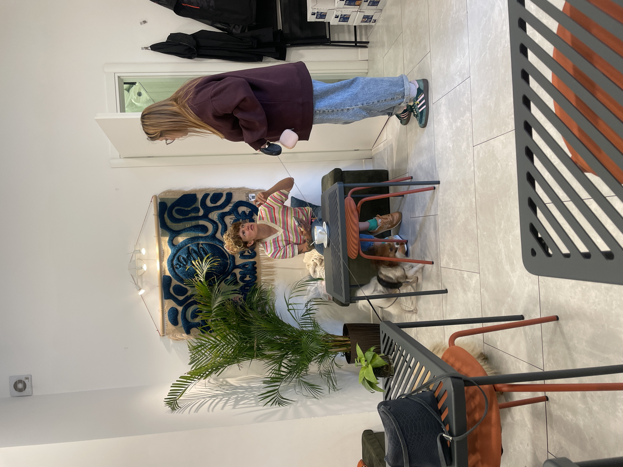 A woman with long blonde hair, wearing a maroon sweater and blue jeans, is facing left and holding a black object in a light-colored store, possibly in Budva, Montenegro. A second woman with curly blonde hair and a striped shirt is seated at a dark table. There are several objects on the table, including a white mug. A dog is lying on the floor near the table. Various plants are visible in the scene. The floor is tiled, and the walls are white. In the background, a blue and white tapestry hangs on the wall. There is a coat rack in the upper left corner of the image. A rack with boxes is visible above the coat rack. Modern chairs are positioned around the table.