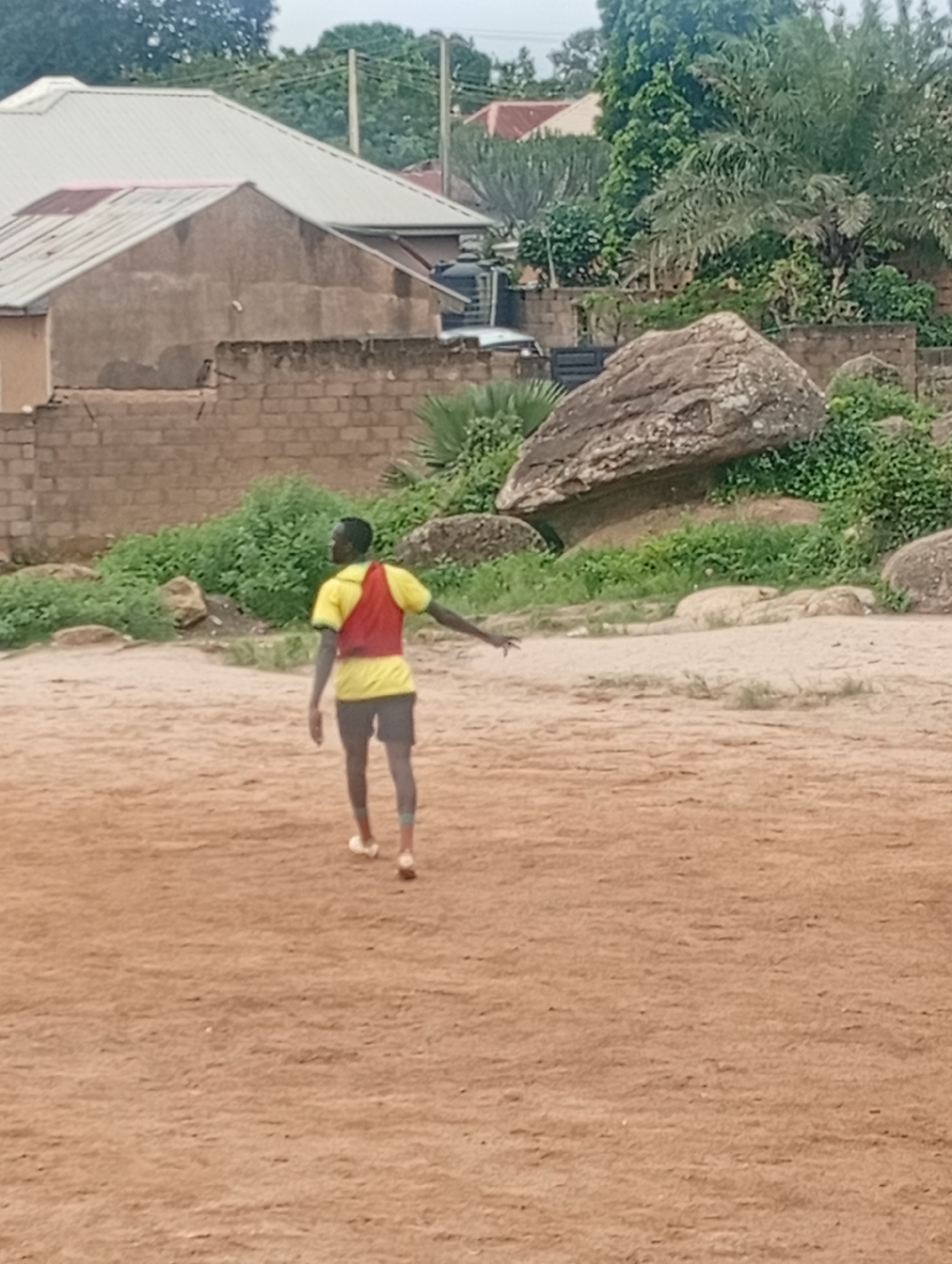 The image depicts an outdoor scene, possibly in a residential area of Jos, Nigeria. In the foreground, a young man wearing a yellow shirt and red vest over dark shorts is walking on a dirt ground. He is facing away from the camera, with his right arm extended. The background features a brick wall, lush green vegetation, and large boulders, including a prominent one in the mid-ground. Behind the vegetation, buildings with corrugated metal roofs are visible, suggesting a semi-urban or developing area. The sky is overcast, indicating a cloudy day. The ground appears to be sandy or dusty, typical of the terrain in parts of Jos.