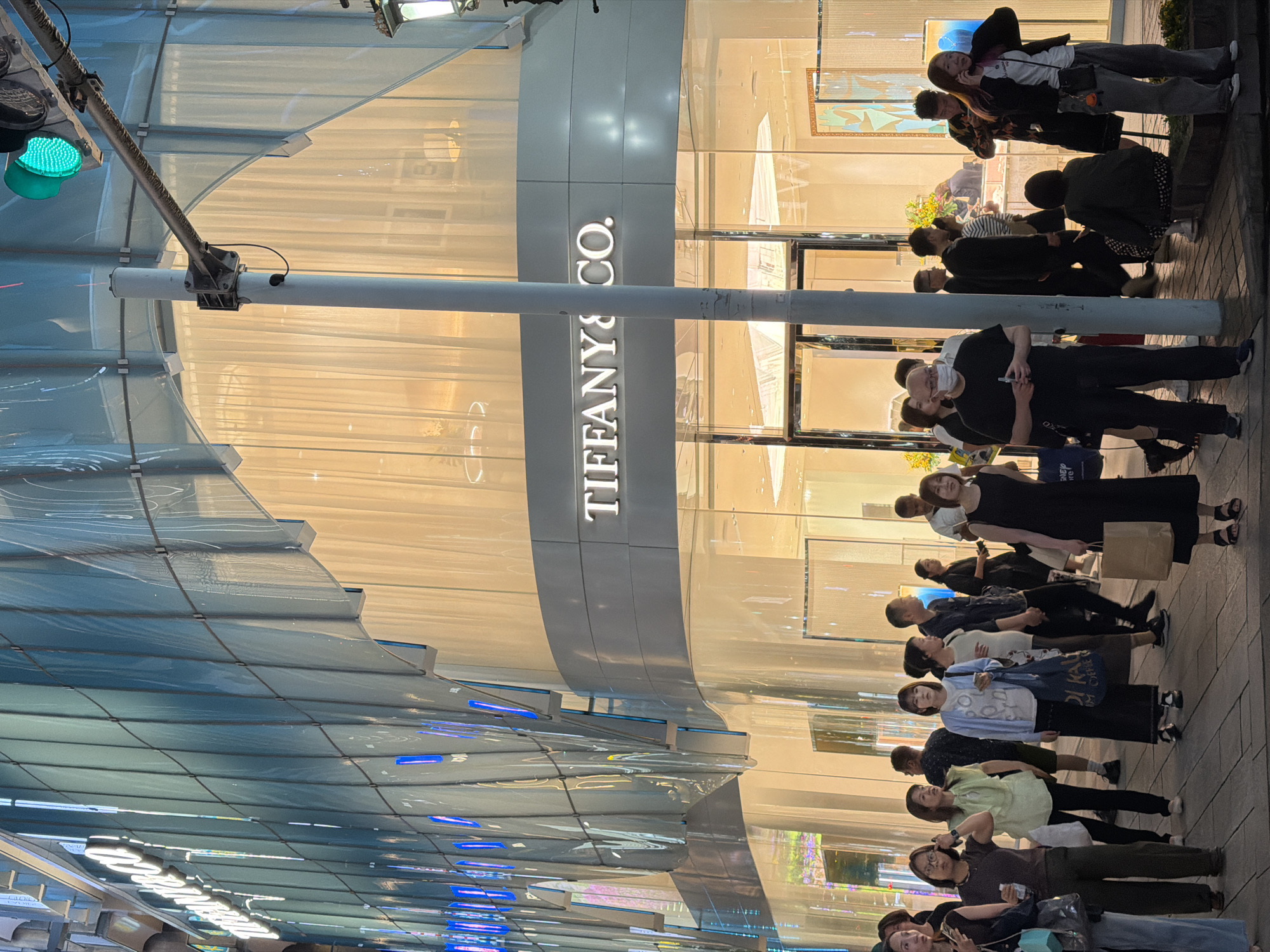 A street-level, medium shot captures the exterior of a Tiffany & Co. store in Chuo, Japan, at dusk. The building's facade is a curved, multi-story structure with a modern design. The upper portion features a translucent, blue-tinted glass curtain wall with vertical elements and integrated blue lighting. Below this, a horizontal band of light-colored, translucent material creates a warm glow, obscuring the interior details but suggesting a retail space. The iconic "TIFFANY & CO." logo is prominently displayed in white, illuminated lettering on a gray band across the lower part of the building. The ground floor features a large glass storefront with entrances and display windows.
In the foreground, a diverse group of people are standing on a paved sidewalk. Their expressions and postures suggest they are either pausing, waiting, or observing. Some individuals are looking at the store, while others are engaged in conversation or simply standing. The lighting is a mix of ambient streetlights, the building's interior and exterior illumination, and a nearby traffic signal that is currently green. A metal pole with attached equipment, possibly a traffic light pole, stands centrally in the image. The overall scene depicts a bustling urban environment in the evening.