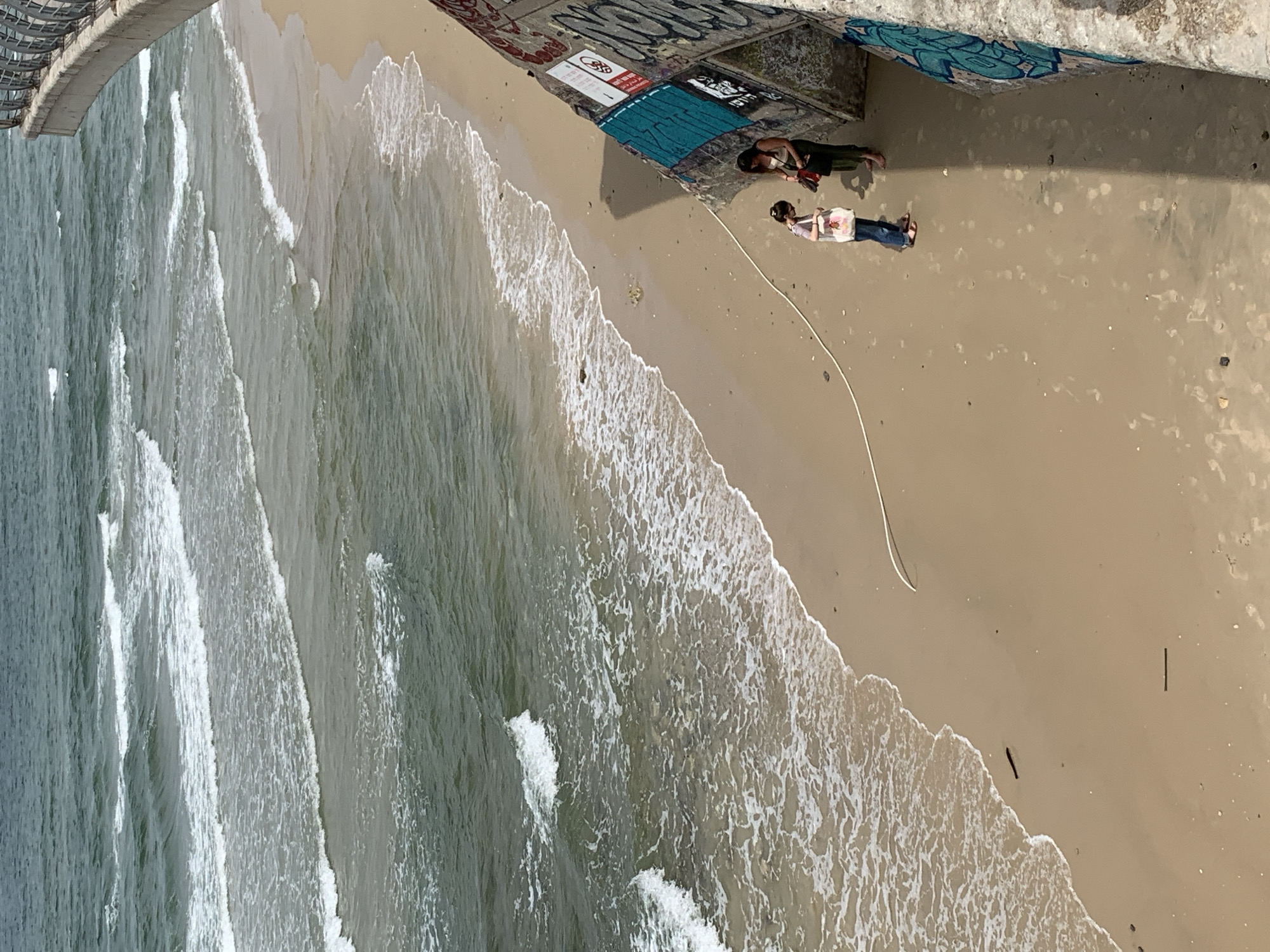 Aerial view of a beach in Tel Aviv, Israel. Two people stand beneath a concrete structure covered in graffiti. One person wears dark clothing while the other wears a white shirt and jeans. A white cable stretches away from them across the sand towards the water. Waves crash on the beach. The scene is lit by bright sunlight.