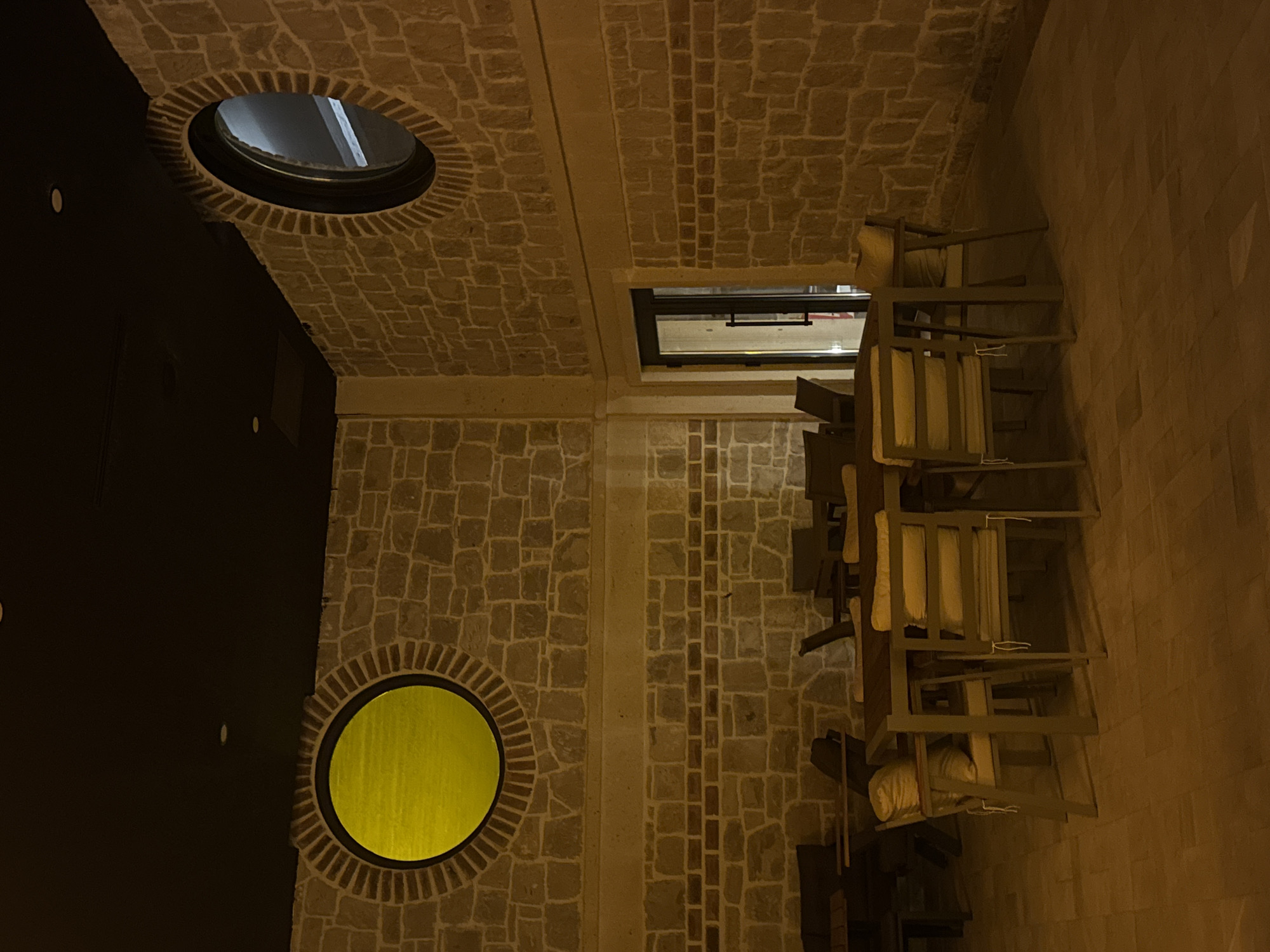 A low-angle indoor shot depicts stacked chairs against a stone wall with a window above, located in Ürgüp, Turkey. The scene is illuminated, revealing details of the building's structure and decor. The chairs, stacked on the right side of the image, feature light-colored cushions. The stone wall is a mix of square and rectangular stones. On the left side of the image, there's a dark wall with round windows, one yellow and one showing sky. The stone patterns, chair placement, and window design are integral to the architectural style of Ürgüp, Turkey. The building appears to be of contemporary design.