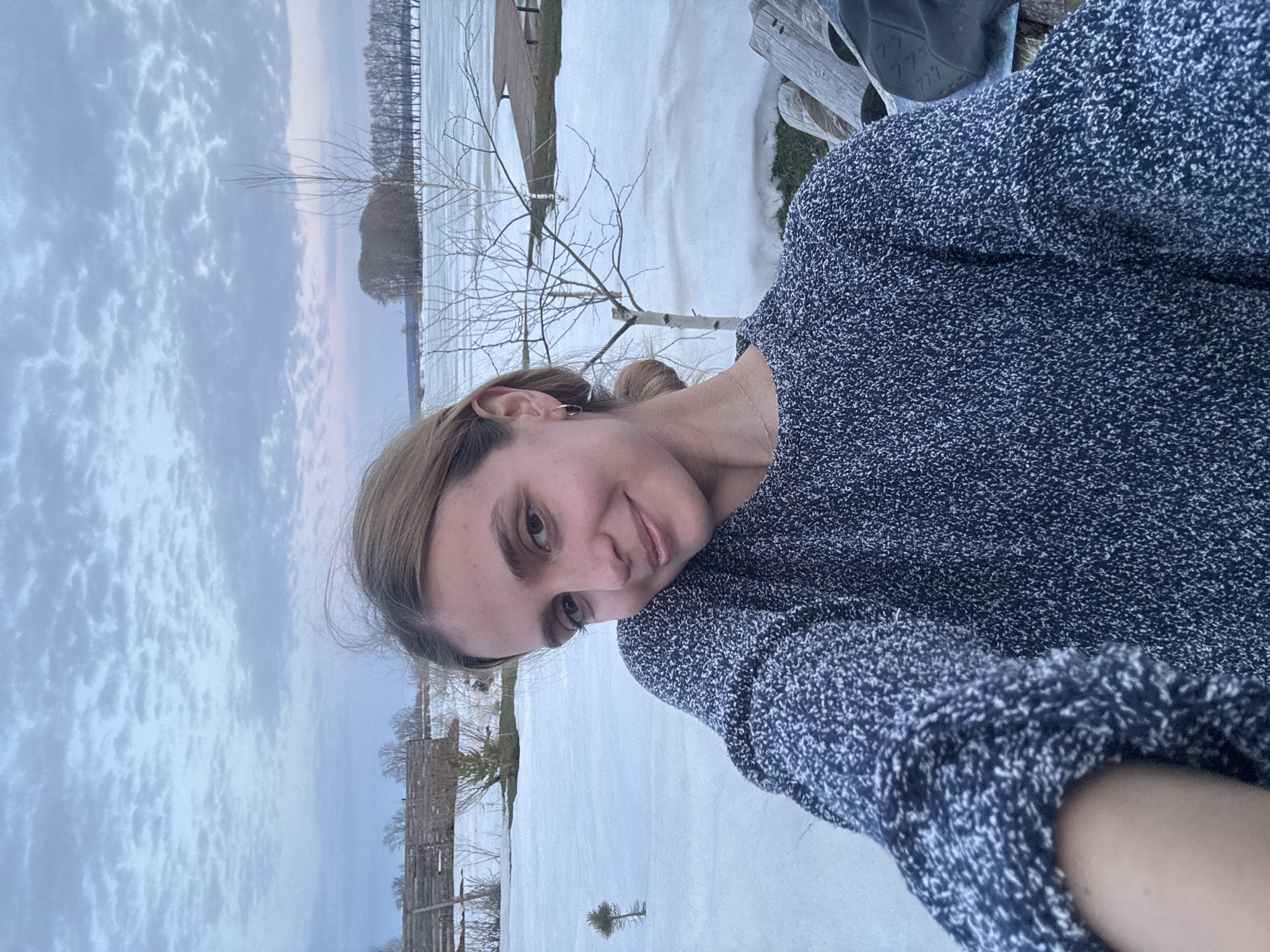 A young adult female subject with light brown hair tied back is depicted in a selfie, smiling slightly and looking directly at the camera. She wears a dark blue and white speckled knit sweater, small silver hoop earrings, and a delicate gold-colored necklace. The background reveals a winter or early spring outdoor environment featuring extensive snow cover on the ground, interspersed with patches of exposed soil or dead grass. A partially frozen body of water, possibly a lake or wide river, is visible, with a wooden dock or platform extending from the right side. A line of bare deciduous trees borders the far bank of the water, and a cluster of trees forms a small island or peninsula further out. In the midground, a bare deciduous tree with light-colored bark stands, alongside a smaller coniferous sapling to the left. A rustic wooden fence or structure is discernible in the left background. The sky above is overcast with textured clouds, displaying subtle shades of blue, grey, and pink/purple, indicating either dawn or dusk. The location is in the city of Unknown, Unknown.