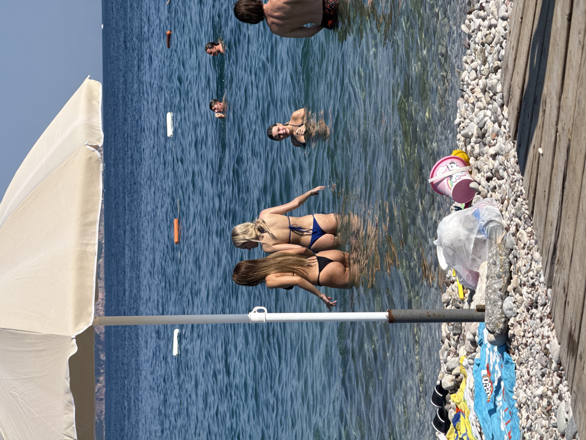 The image depicts a sunny day at a pebble beach in Budva, Montenegro. In the foreground, a white beach umbrella provides shade, and its pole stands in the shallow water. The shoreline is covered in smooth, grey and white pebbles, with a wooden walkway visible on the right. Several beach towels and a child's pink bucket and spade are scattered on the pebbles.
In the water, two women in bikinis are standing, facing away from the camera, with the sea reaching their waists. To their right, a young woman in a bikini is partially submerged, looking towards the camera. Further out in the blue sea, several other individuals are visible, some with their heads above water. Two white buoys and an orange buoy are floating in the water, marking a designated area. The background shows a clear blue sky and the silhouette of a distant coastline.
