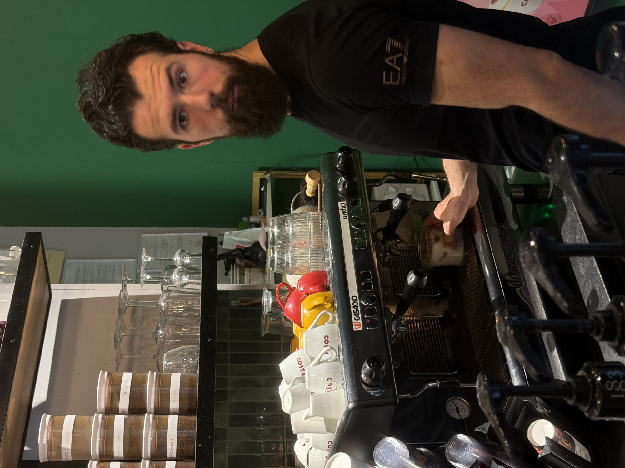 A man with a beard stands behind a coffee machine, seemingly making coffee. He wears a black t-shirt with the letters "EA" visible on the shoulder. The coffee machine, labeled "Undici" and "Casadio," is surrounded by stacked Costa coffee cups, red and yellow mugs, and various glasses on a metal shelf. Spice jars with labels like "Kenya," "Corcovado," "Mactan," and "Quaten" are also visible on the shelf. The background is a green wall. The location is Podgorica, Montenegro.