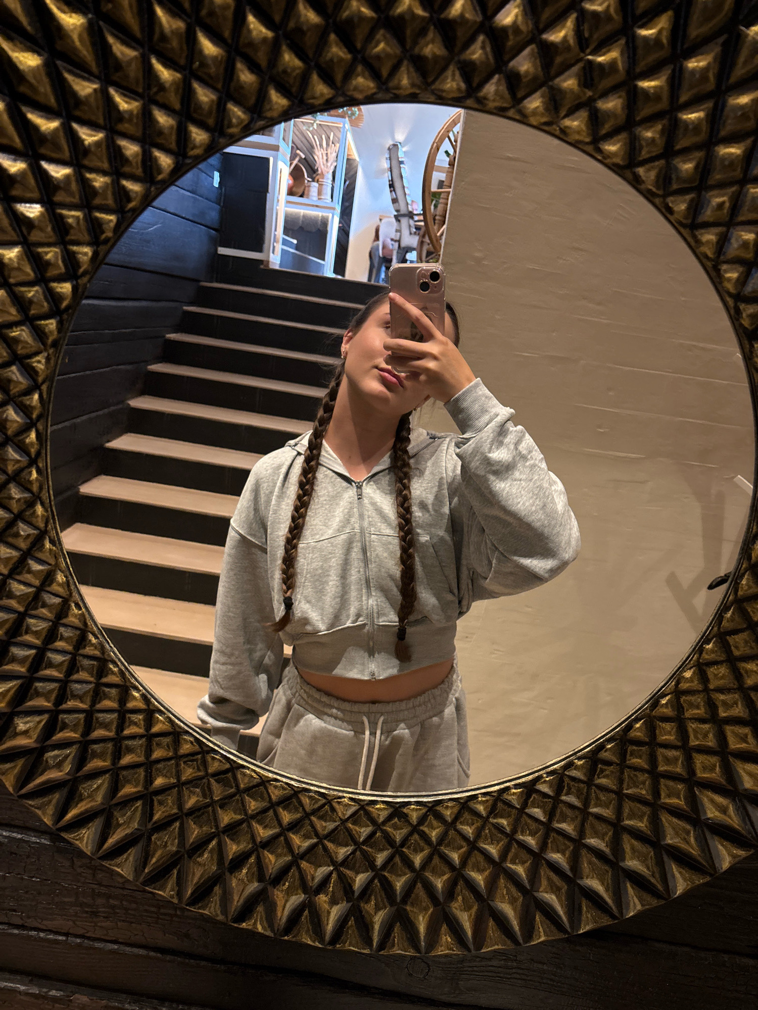 A woman with braided hair is taking a mirror selfie. She is wearing a grey cropped hoodie and grey sweatpants. The mirror she is using has an ornate gold frame with a diamond pattern. In the reflection of the mirror, a staircase with dark wooden steps and black risers is visible. Beyond the staircase, there is a display of decorative items on shelves. The setting appears to be an indoor location, possibly a hotel or a stylish residential space, in Vorokhta, Ukraine.