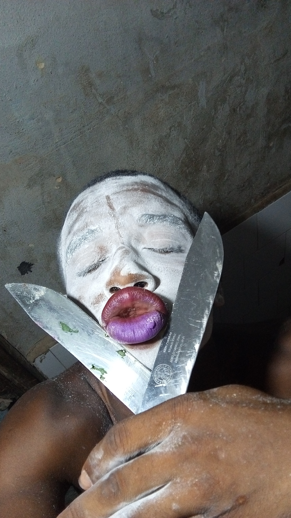 The image captures a person, likely male, with dark skin, extensively covered in white powder from their forehead to their neck and visible hands. Their eyes are closed, and their lips are dramatically exaggerated, appearing swollen and colored a deep purple mixed with red. Two large, shiny silver knives are prominently positioned in front of the person's face, crossing each other in an 'X' shape. Small green specks, possibly herbs or food residue, are visible on the blade of the left knife. A hand, also dusted with white powder, is visible in the lower right foreground, seemingly holding one of the knives.
The setting appears to be indoors against a dark, textured background that resembles a concrete or unfinished wall/ceiling. The lighting is dim and casts shadows, contributing to a stark and somewhat dramatic atmosphere. The overall impression is that of a performance, a costume, or a ritualistic depiction, evoking a sense of the surreal or unsettling.
On the blade of the right knife, faint and mirrored text can be observed, including what appears to be the word "STEEL" along with other illegible characters, likely part of a manufacturer's inscription. The exact time of day and specific location are not discernible from the image.