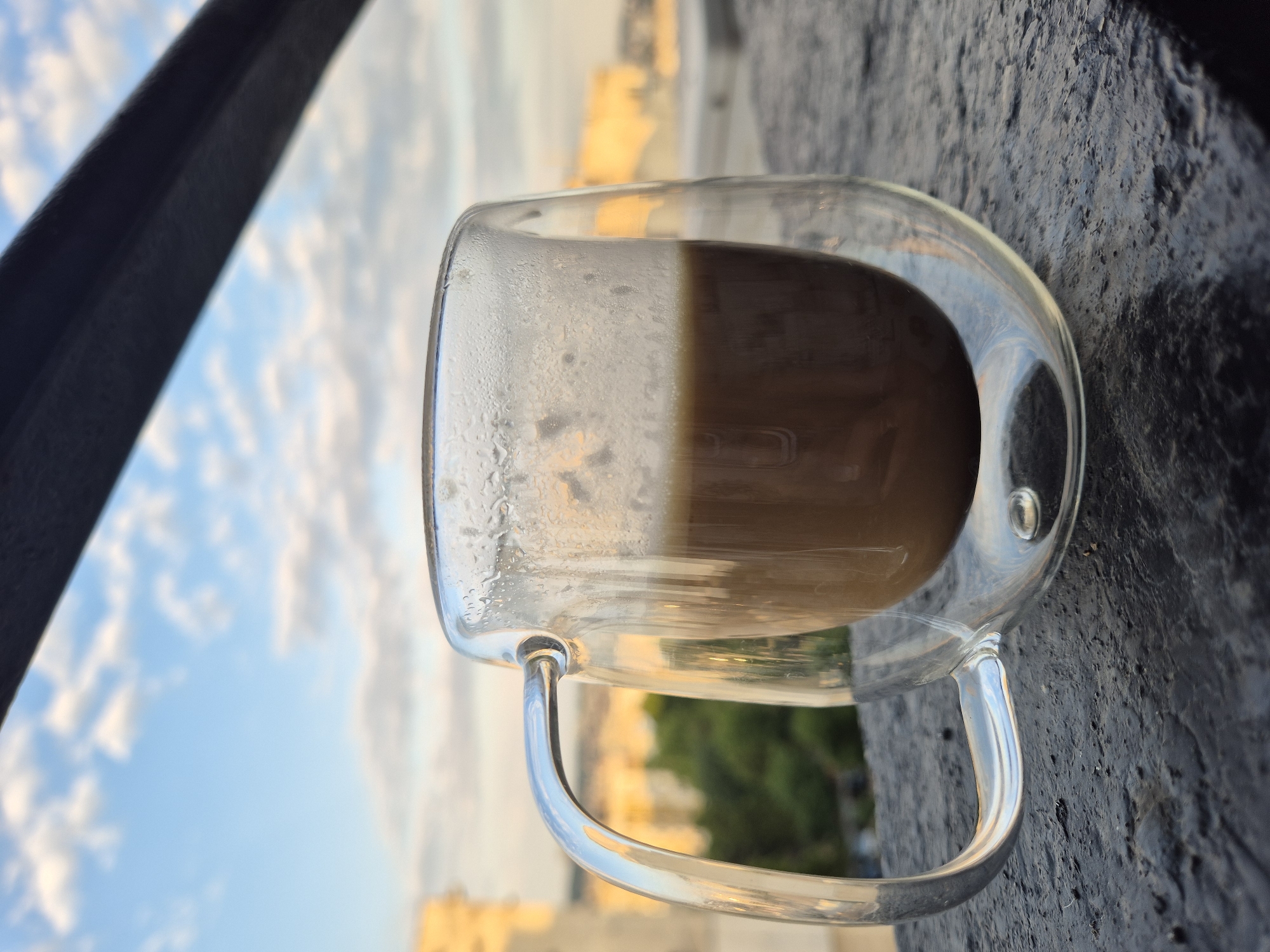 A close-up shot focuses on a double-walled glass mug filled with coffee, positioned on a dark, textured surface. The mug's handle is prominently displayed on the left, reflecting the sky. The background features a blurred cityscape with buildings bathed in warm sunlight and a sky filled with scattered clouds. The overall atmosphere suggests a peaceful morning or evening setting, possibly from a balcony or rooftop overlooking the city.