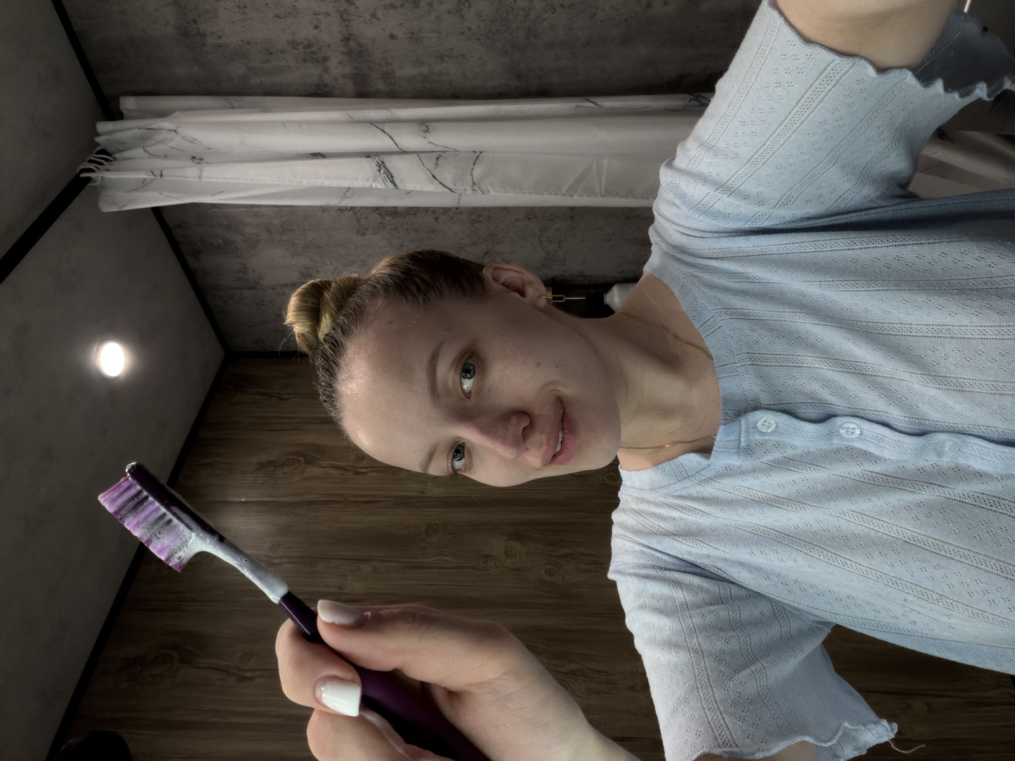 A woman is holding a toothbrush with toothpaste in what appears to be a bathroom. The woman has her blonde hair up in a bun. She is wearing a light blue button-up top. The bathroom has a shower curtain, and a light fixture is visible. There is no information that indicates the location of this image is Reshetnikovo, Russia.