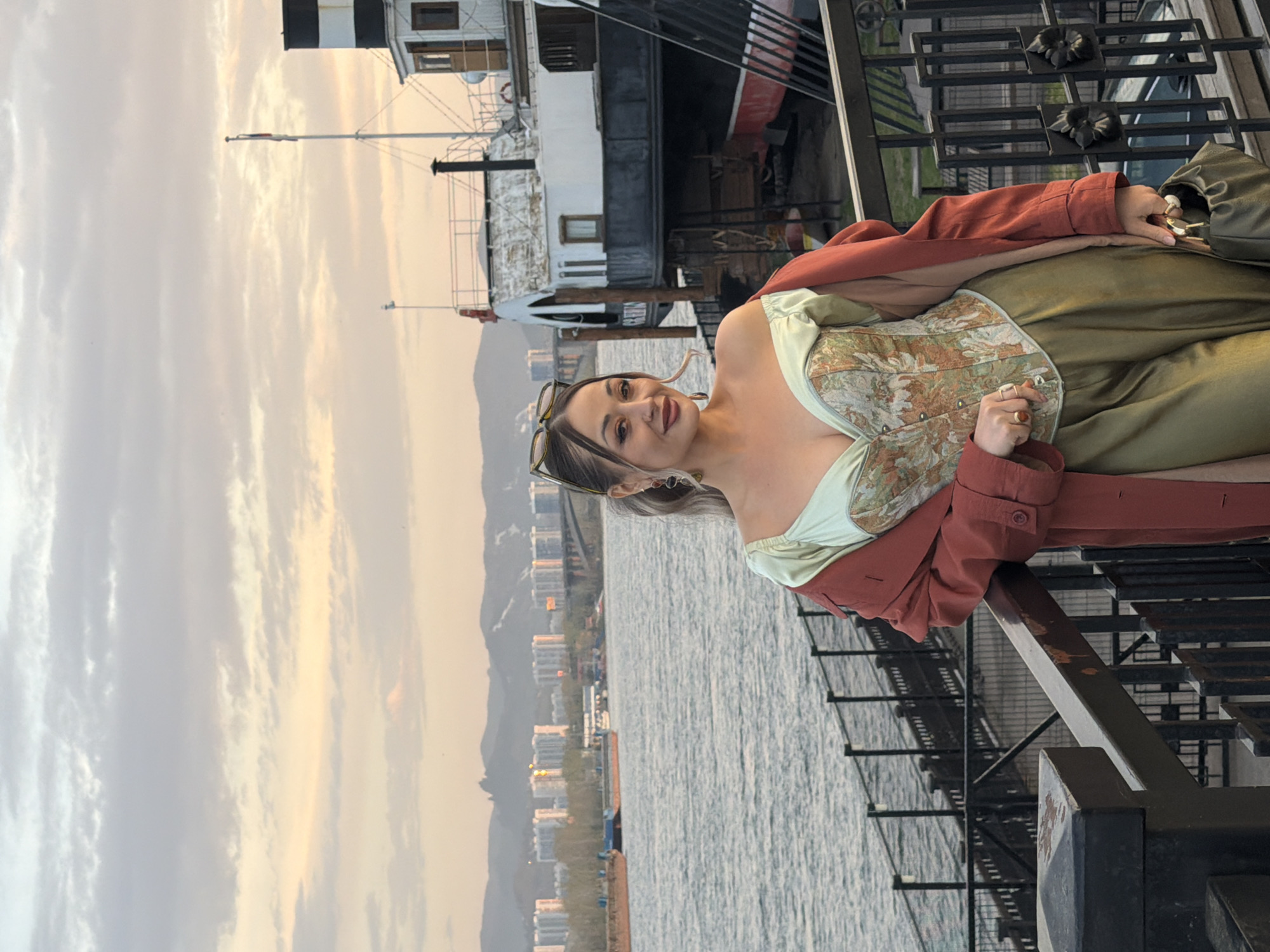 A young woman sits on a black metal railing by the Yenisey River in Krasnoyarsk, Russia. She wears a green corset top, a red jacket, and olive-colored pants. She holds a green purse. The river is in the background, along with buildings and mountains obscured by haze. A ship-like structure with black and white stripes can be seen behind her. The sky is cloudy and has an orange hue from the sunset.