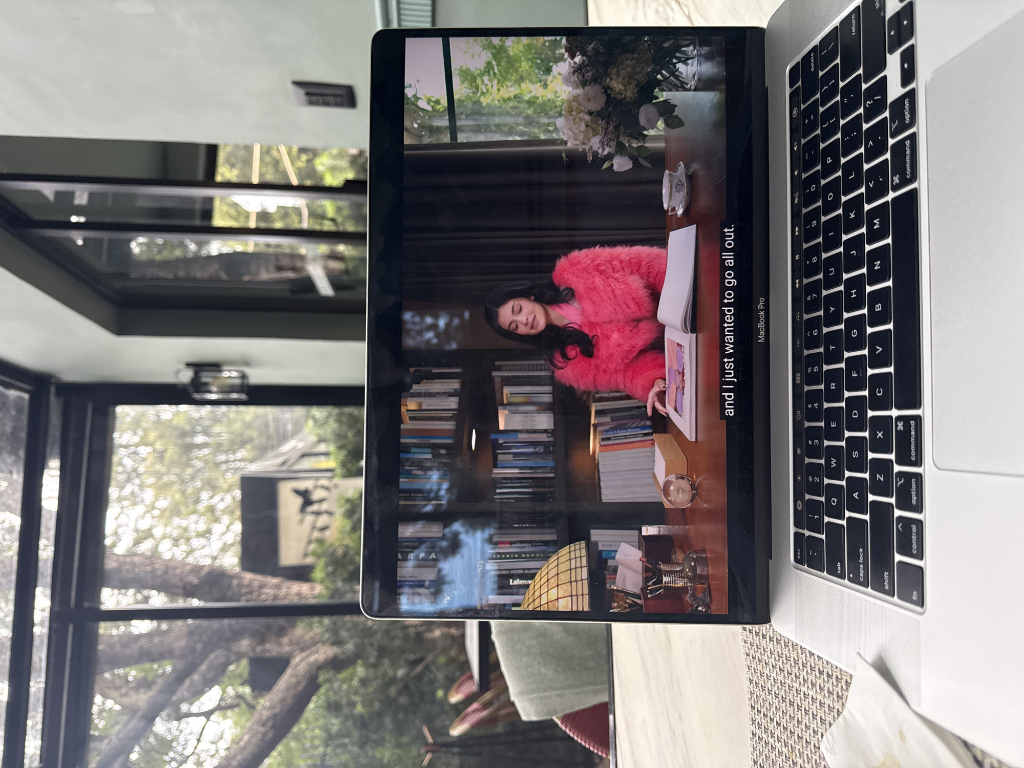 The image shows a laptop open on a table, displaying a scene from a video. The video features a young woman with dark hair wearing a bright pink fluffy jacket, sitting at a desk with a book open in front of her. Behind her is a bookshelf filled with books. To the right of the woman on the desk is a vase with flowers and a teacup and saucer. The laptop is a MacBook Pro.
The scene is viewed from behind the laptop, looking towards the laptop screen. The laptop is placed on a textured placemat, which rests on a marble-topped table. In the background, through a large window or glass wall, trees and outdoor seating areas are visible. The overall environment appears to be an indoor space with a view of a garden or patio, possibly a cafe, restaurant, or a sunroom. There is no specific indication of the location being in Mexico City, Mexico.