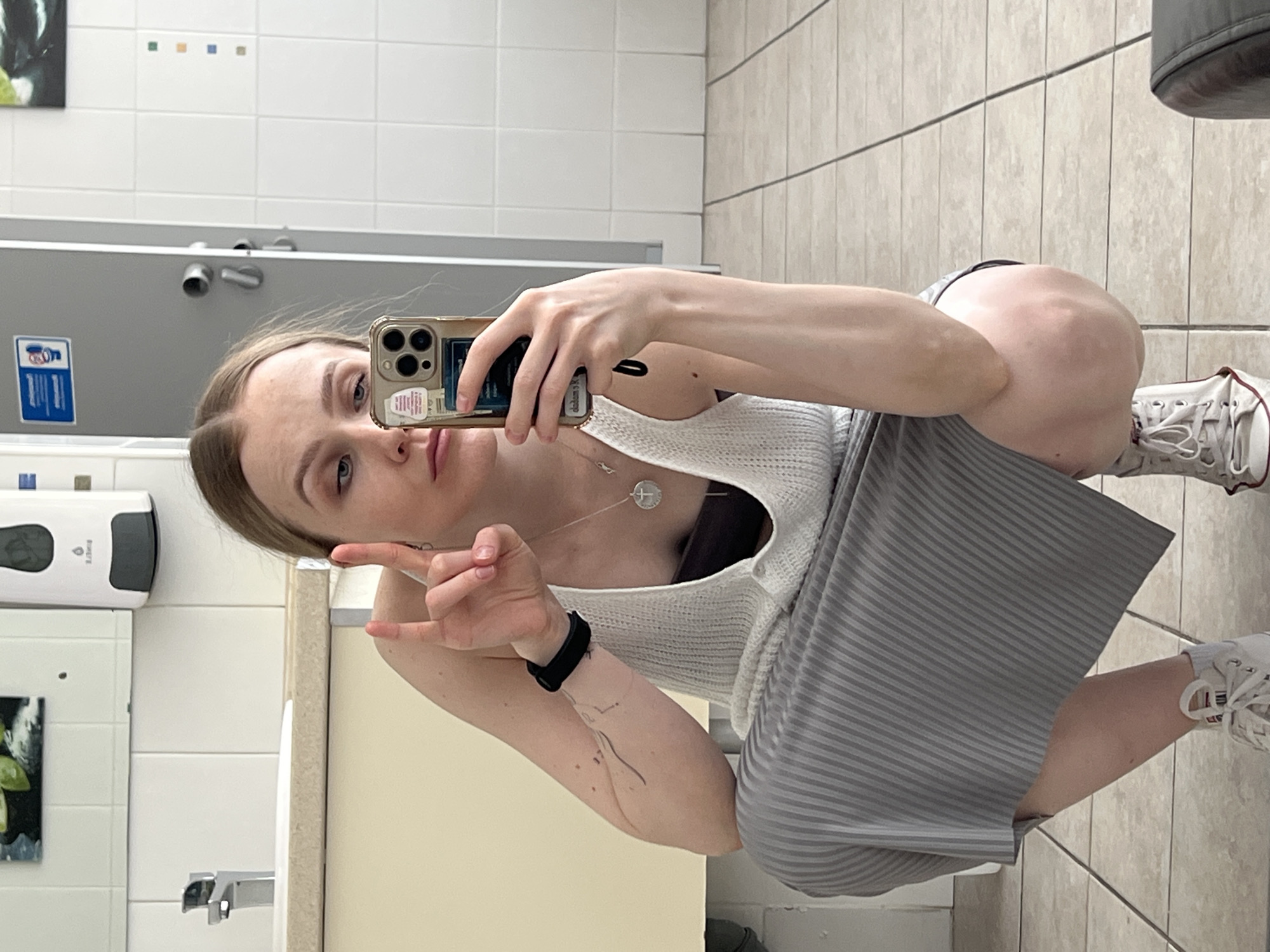 A young woman squats in a restroom while taking a selfie with a smartphone. The restroom is clean, with white and gray tiled walls and floors. The background includes a sink, soap dispenser, and a metallic toilet stall divider. The woman is wearing a white tank top, a gray striped skirt, and white sneakers. She is making a hand gesture toward the camera. The location is presumably in a public space in Khimki, Russia.