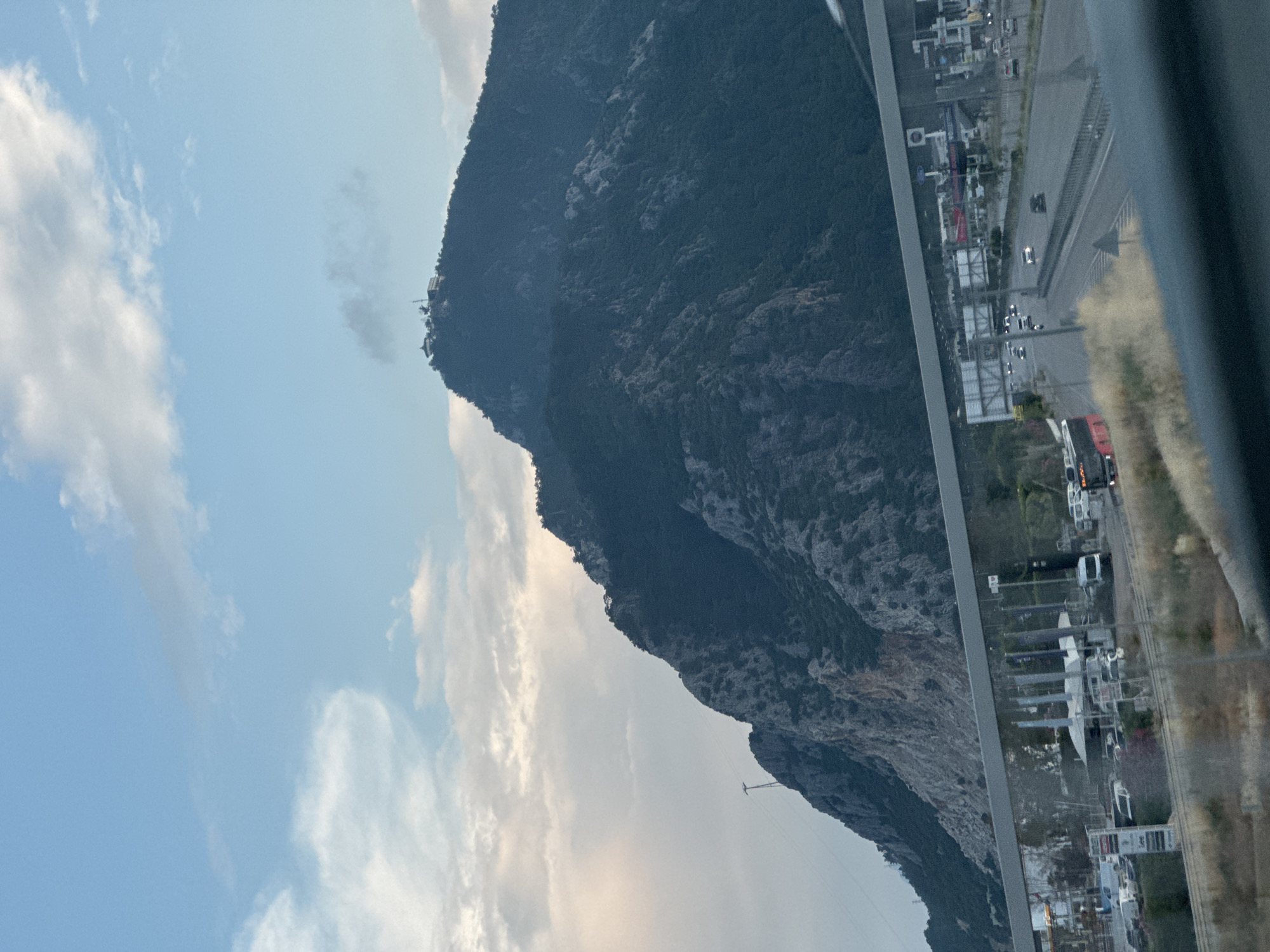 The image shows a view from inside a building or vehicle, through a glass panel. The foreground contains a portion of the building interior, blurred and out of focus. Beyond the glass, the landscape includes a mountain with rocky terrain covered in vegetation. The sky is partly cloudy, with shades of blue and white. A structure is visible near the mountain peak. To the right, a section of a highway is seen with vehicles. Adjacent to the highway, an urban area is visible with buildings and commercial establishments. Based on the location, it is likely near Konyaaltı, Turkey.