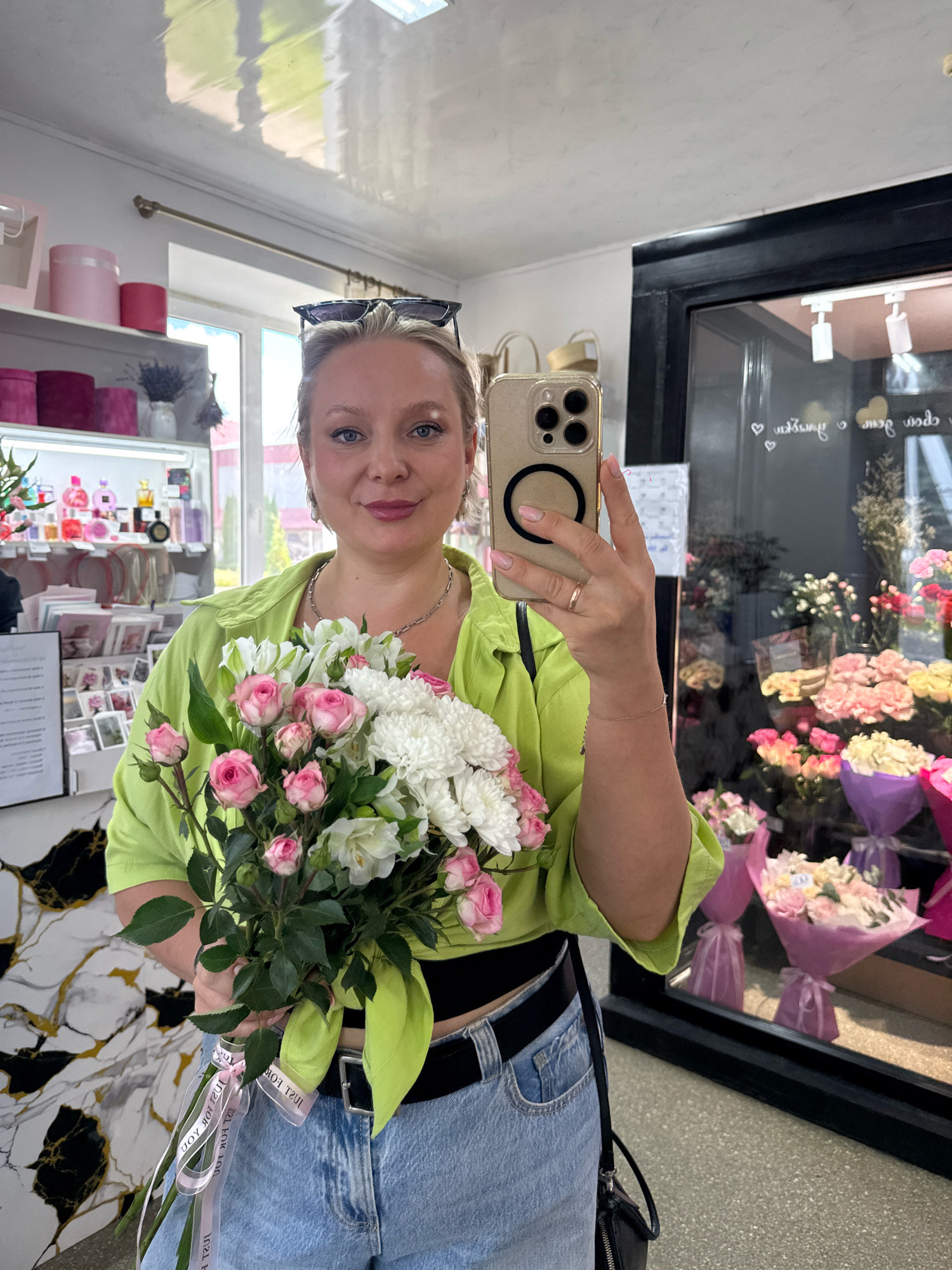 The image shows a woman taking a selfie in a flower shop, likely in Slawharad, Belarus, based on the context. She is standing in front of a display of flowers. The woman is holding a bouquet of pink roses and white chrysanthemums. She is wearing a lime green shirt and blue jeans. Her hair is blonde and she is wearing sunglasses on top of her head. The flower shop appears to be well-stocked with a variety of flowers and floral arrangements.