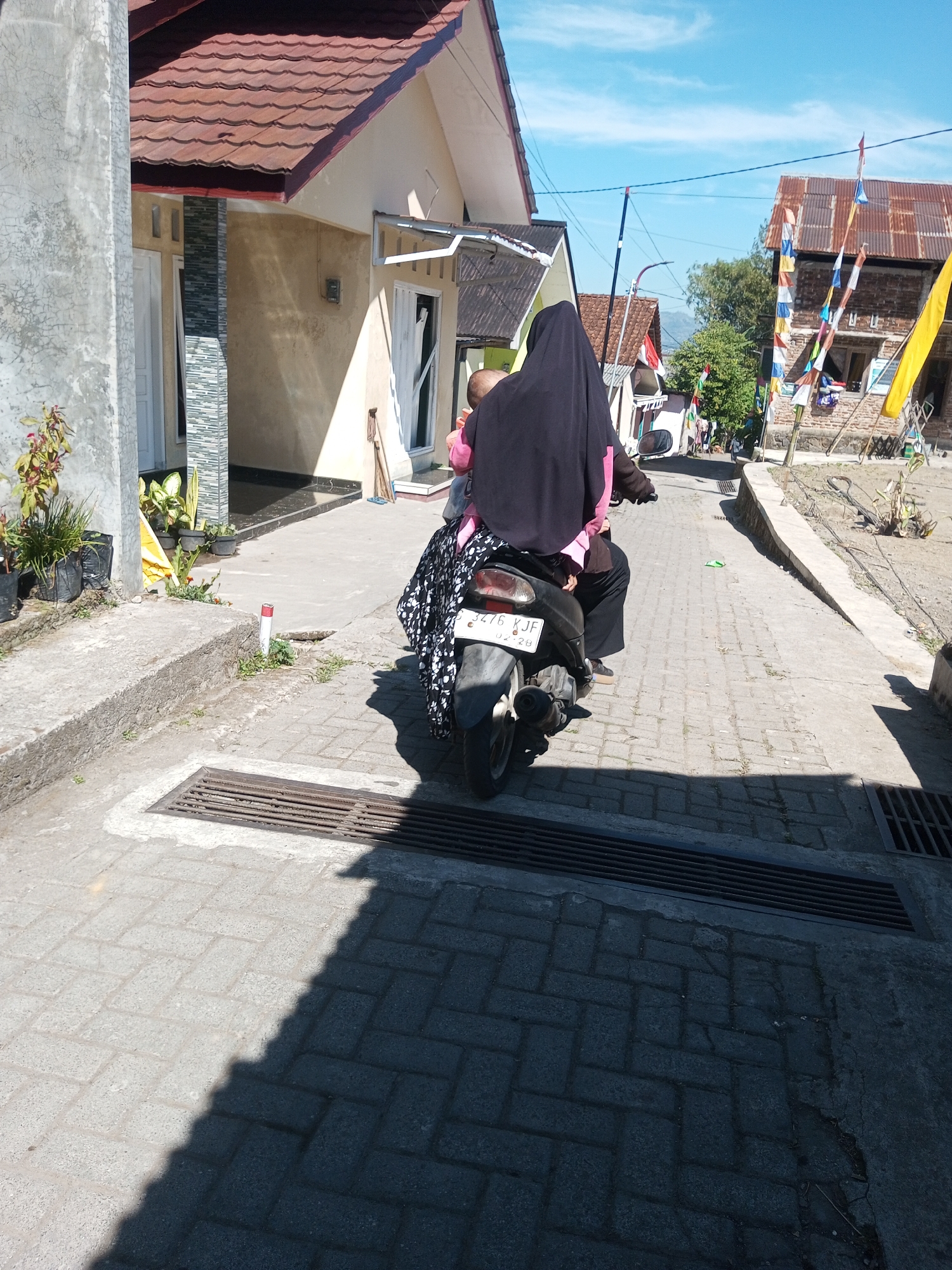 The image depicts a street scene in Andongsili, Indonesia, bathed in bright sunlight. A person is riding a scooter with another person as a passenger, both wearing traditional Indonesian attire. The passenger is wearing a black hijab and a pink veil. The scooter is covered with a patterned cloth. The street is paved with interlocking bricks, and a drain grate is visible in the foreground. To the left, a house with a red tiled roof and white walls is partially visible. The background shows more houses, some adorned with colorful flags, and lush green trees. The sky is a clear, vibrant blue.