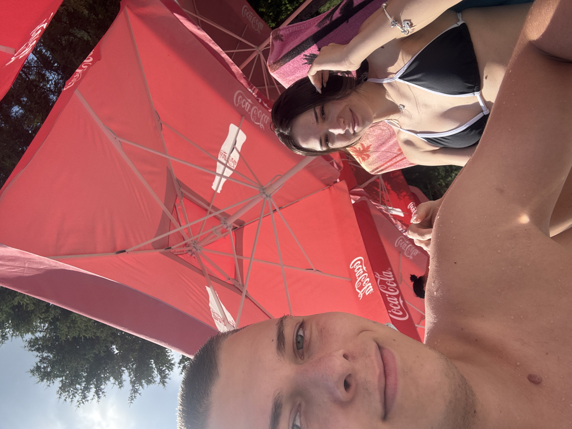The image shows a young couple in Chiflik, Bulgaria, sitting under red umbrellas. In the foreground, a young man with short dark hair and green eyes is smiling at the camera. He is shirtless, and his skin appears to be wet, possibly from swimming. To his right, a young woman with long dark hair is wearing a black bikini top with white trim. She is looking at the camera and smiling, with one hand touching her hair. She is wearing a necklace and a bracelet. Both individuals are seated, and the background features red umbrellas with Coca-Cola branding, trees, and a bright sky. The overall scene suggests a relaxed day in an outdoor setting, likely a resort or recreational area.