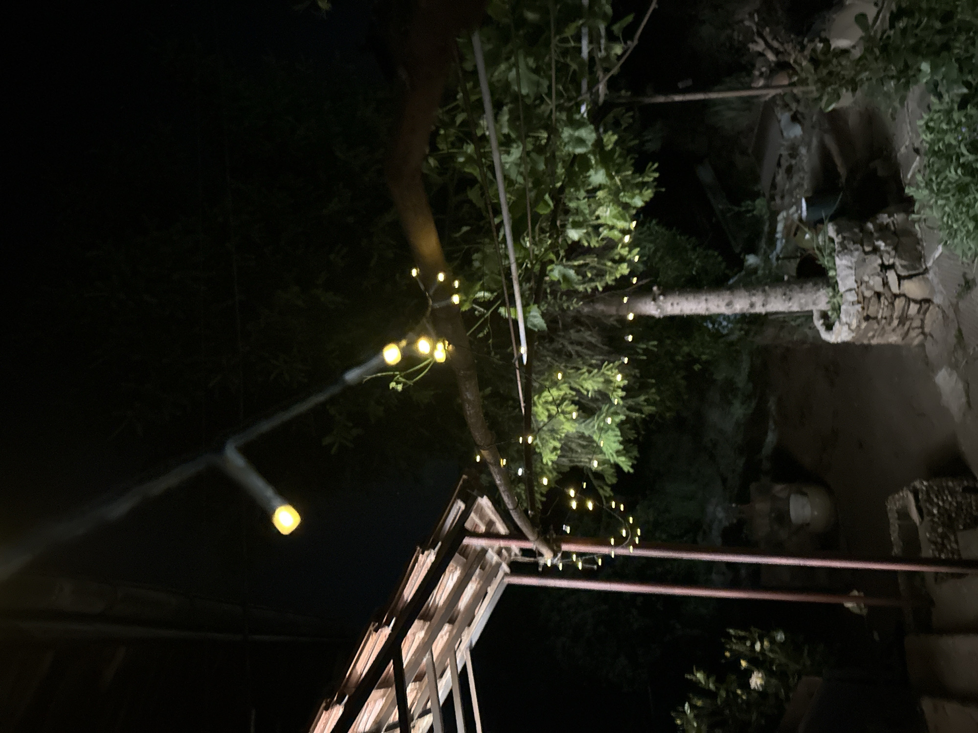At night, a view is seen from beneath a wooden structure, possibly a pergola or arbor. The structure consists of regularly spaced planks spanning between heavier beams. String lights are wrapped around a tree and parts of the structure. In the upper right, a stone wall, vegetation, and stairs lead to a higher level. A red metal rail is present. The area is dimly lit, with most light emanating from the string lights. The location is near Slavyani, Bulgaria.