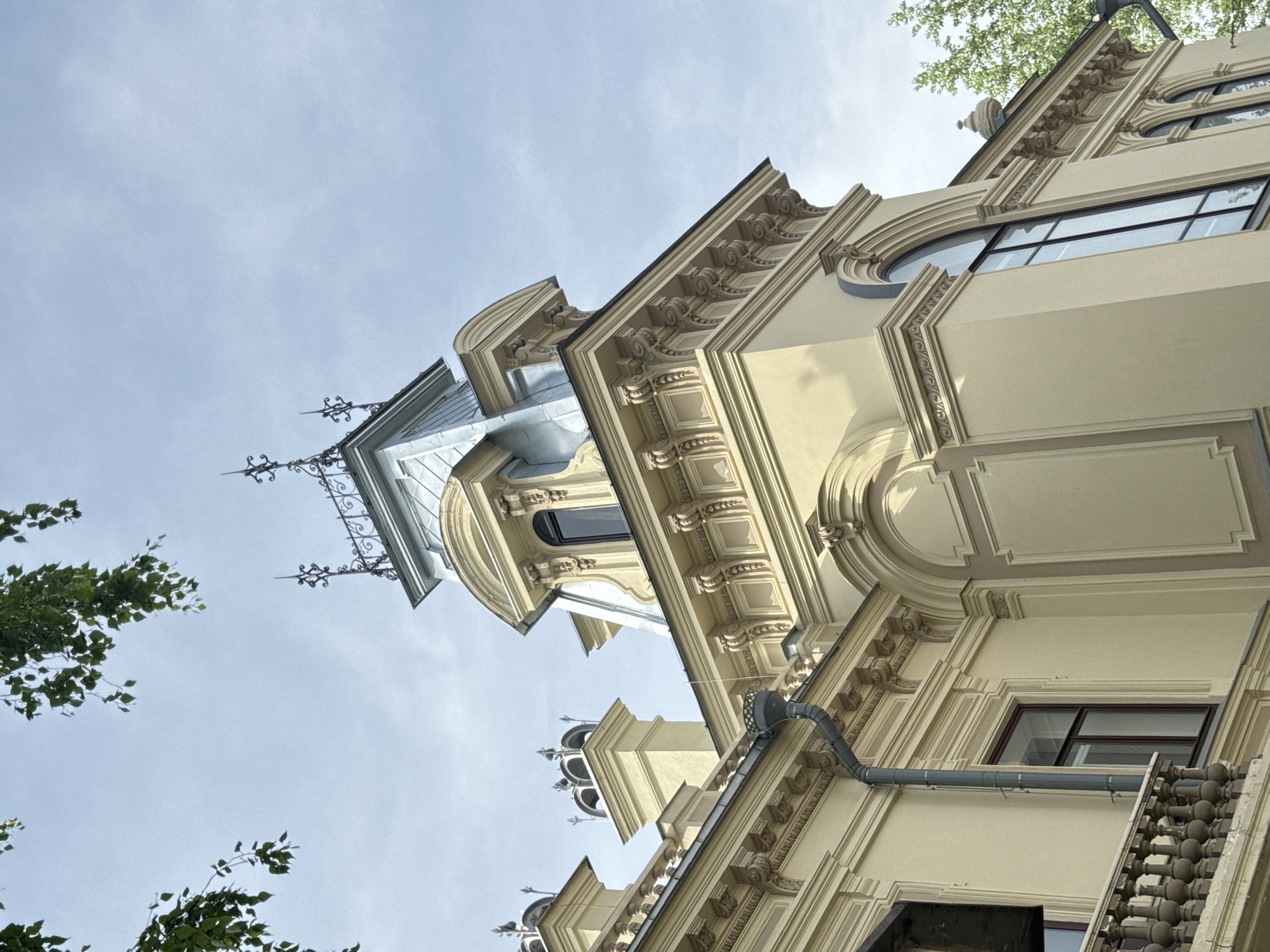 The image depicts a low-angle view of a building facade in Kazan, Russia. The architectural style is ornate, featuring detailed moldings, decorative cornices, and a turret-like structure with a metal roof and wrought-iron details. Windows with dark frames are visible, and a gray drainpipe runs vertically along the wall. Branches with green leaves are present in the lower left corner, suggesting nearby trees. The sky is overcast, with a light blue-gray hue.
