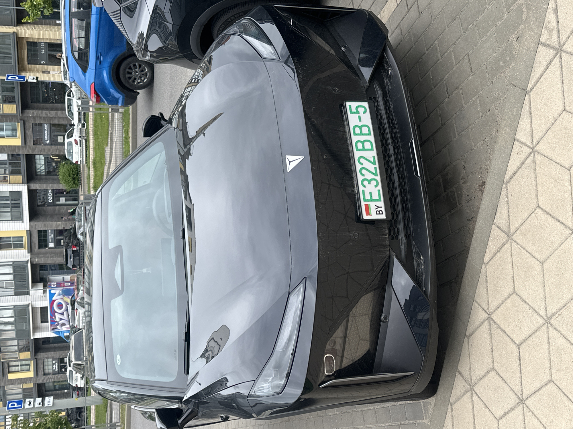 The image shows the front of a black car with a Belarusian license plate (E322BB-5) parked on a paved surface in Бараўлянскі сельскі Савет, Belarus. The car's license plate has the country code "BY" at the bottom. The car is next to another car (partially visible in blue) and several other cars parked along the street. Buildings, including one with an "OZON" sign, are visible in the background.