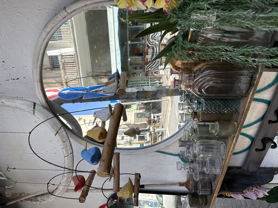 The image displays a detailed view of an outdoor storefront display, featuring a large, round mirror reflecting a street scene in Tel-Aviv, Israel.
In the foreground, a decorative mobile consisting of multiple colorful, stylized wooden birds is suspended. These birds, rendered in red, blue, yellow, and white, are perched on short, horizontal wooden dowels, which are in turn suspended by thin black wires from an unseen structure above. Below this mobile, a prominent round mirror with a distressed white, possibly rattan or wicker, frame is mounted against a white stucco wall that shows faint traces of green painted patterns or graffiti.
Immediately beneath the mirror, a wooden shelf holds a collection of approximately 25-30 glass bottles and vases of varying sizes, shapes, and hues. These include clear, amber, light purple, and green glass, with some pieces featuring ribbed or textured designs. To the right of the shelf, a planter contains artificial green trailing foliage and several yellow, orchid-like flowers. Below the shelf, a dark wooden panel with two dark, decorative hooks is affixed to the wall.
The mirror's reflection captures a street in Tel-Aviv. Visible elements include a multi-story building with a light-colored facade and an upper-level balcony featuring a white railing. A blue circular Israeli traffic sign indicating a mandatory right turn, alongside another blue rectangular sign, is prominently reflected on a street pole. Further down the reflected street, commercial establishments are visible: an area with white outdoor tables and chairs to the left, and a closer building with a light blue awning, an interior counter, and a distinctive black-and-white striped wall or mural. A bicycle is partially visible near this establishment. The scene is illuminated by natural light, indicating a clear day.