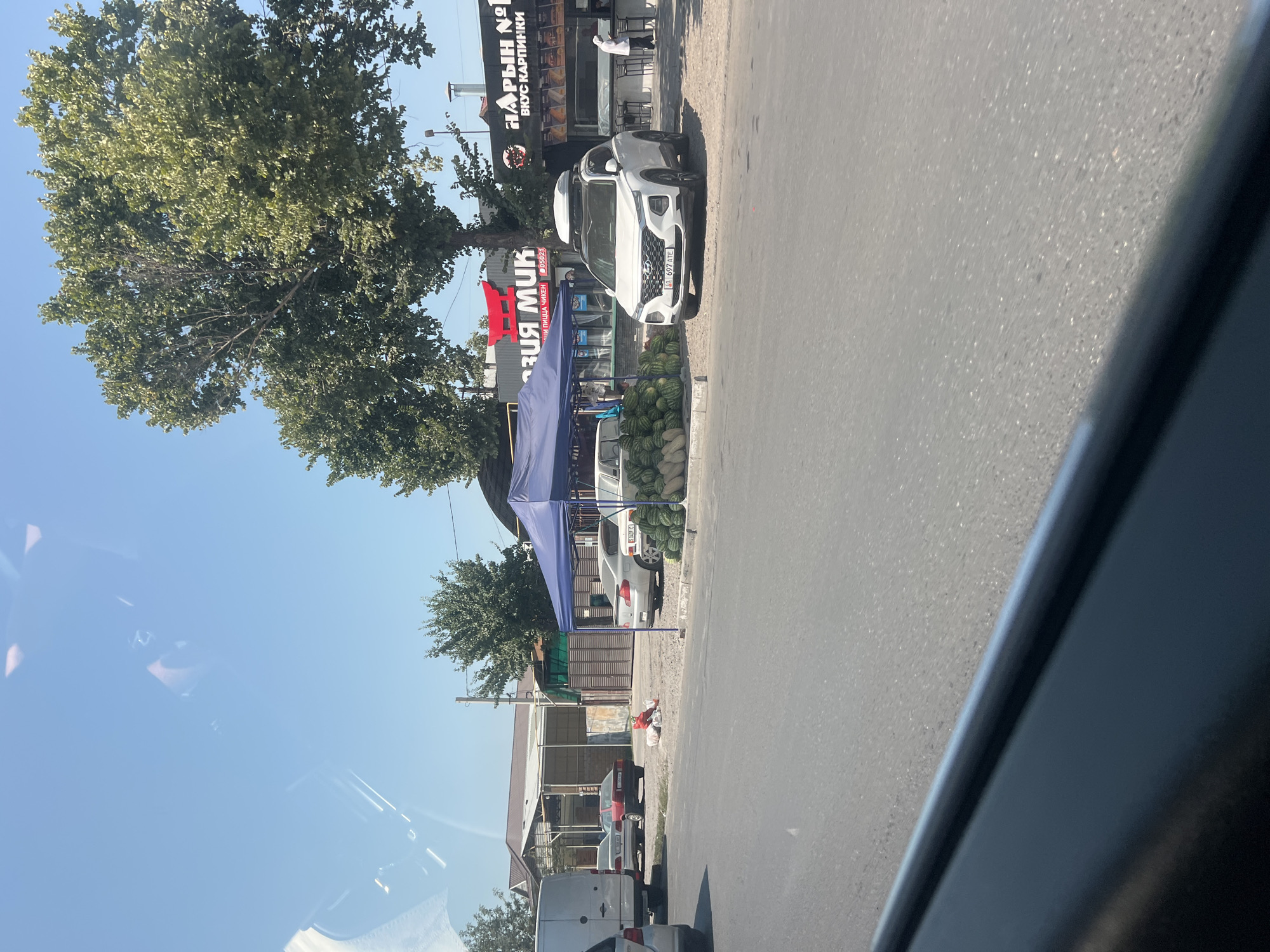 A street scene in Bishkek, Kyrgyzstan, depicts a roadside market on a sunny day. A blue tent is set up, displaying a large pile of watermelons. Several cars are parked nearby, including a white SUV with a roof carrier. Behind the market, buildings with signs, one reading "АЗИЯ МУК", are visible, along with trees and utility poles. A person in a white garment is seen near one of the buildings. The foreground shows the road surface and a dark car interior, indicating the image was taken from a vehicle.